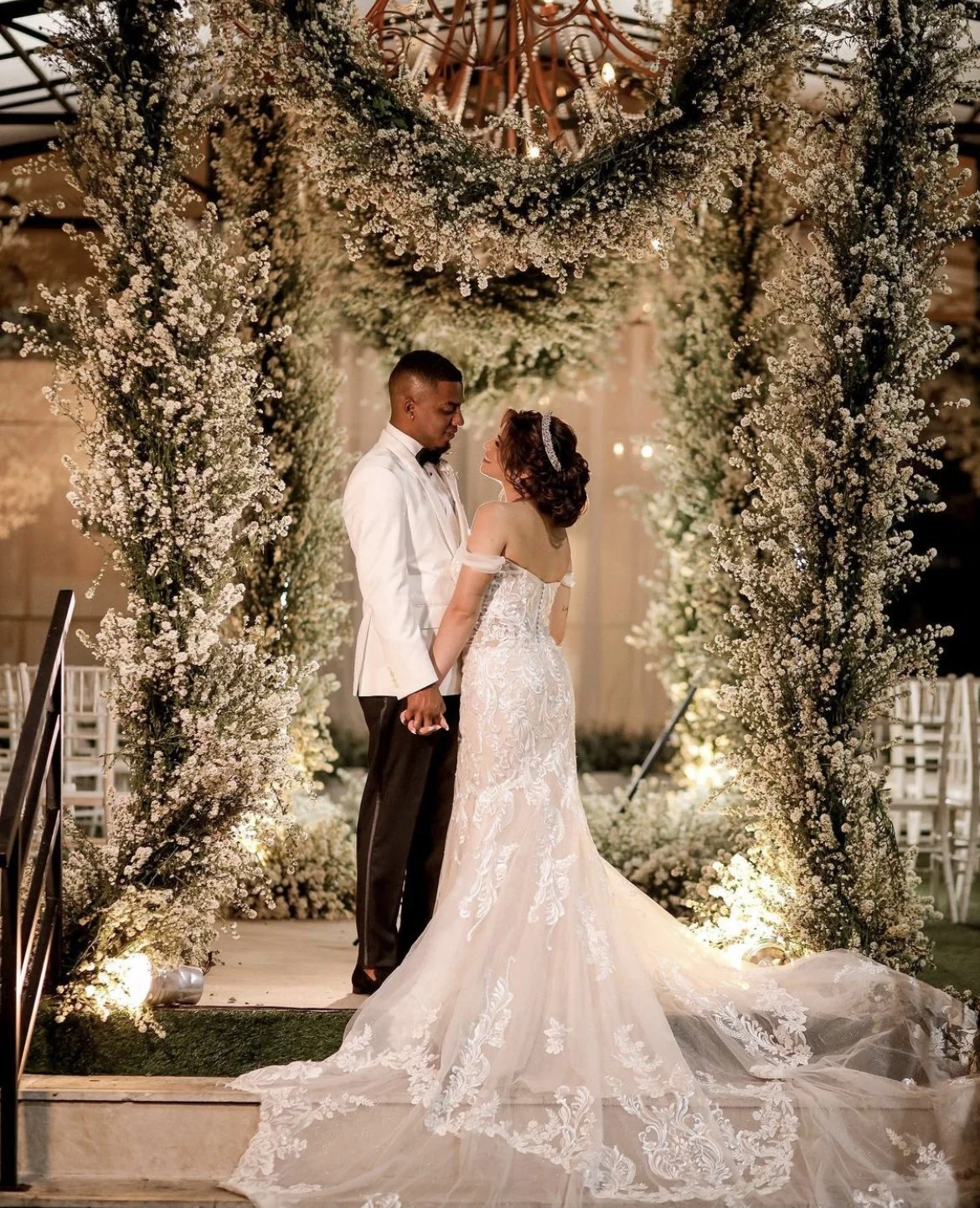 A bride and groom standing under a floral arch during their wedding ceremony, holding hands and gazing at each other.
