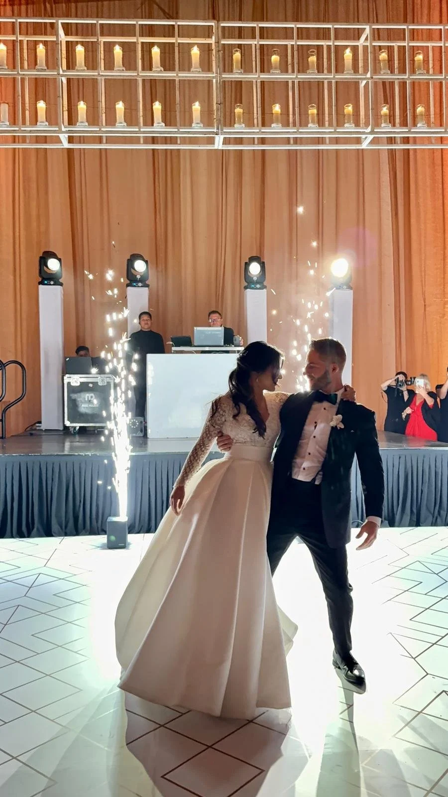 A bride and groom dancing on the dance floor during their wedding reception, with a DJ and guests in the background, and sparklers on the floor.