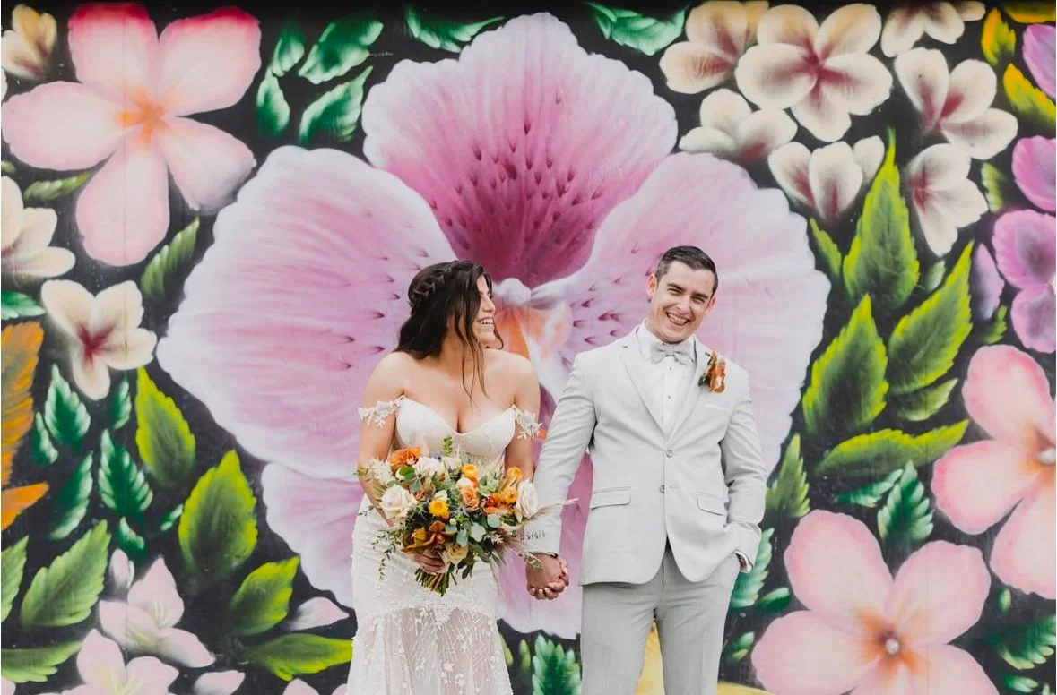 A bride and groom holding hands and smiling, standing in front of a large floral mural with pink, white, and green flowers.