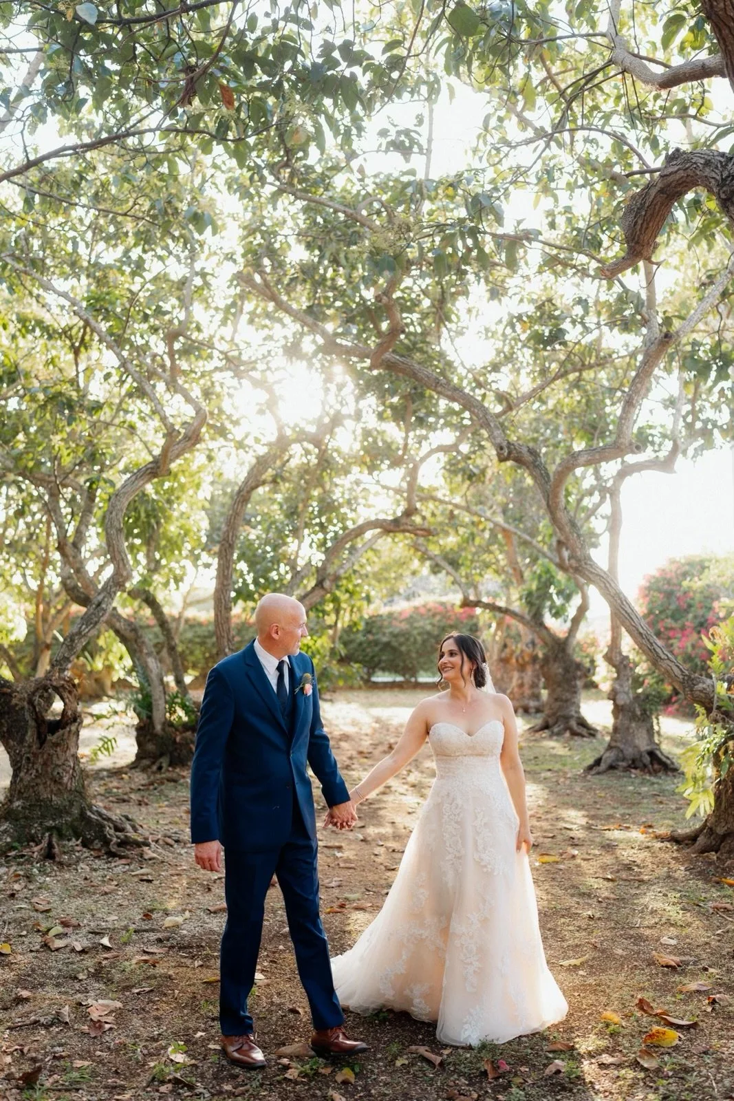 Bride and groom holding hands and smiling in a wooded outdoor setting during sunset.