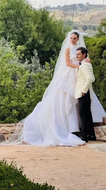 Bride and groom standing outdoors on a dirt path, surrounded by greenery, with the bride in a white wedding gown and veil, and the groom in a white suit jacket and black pants.