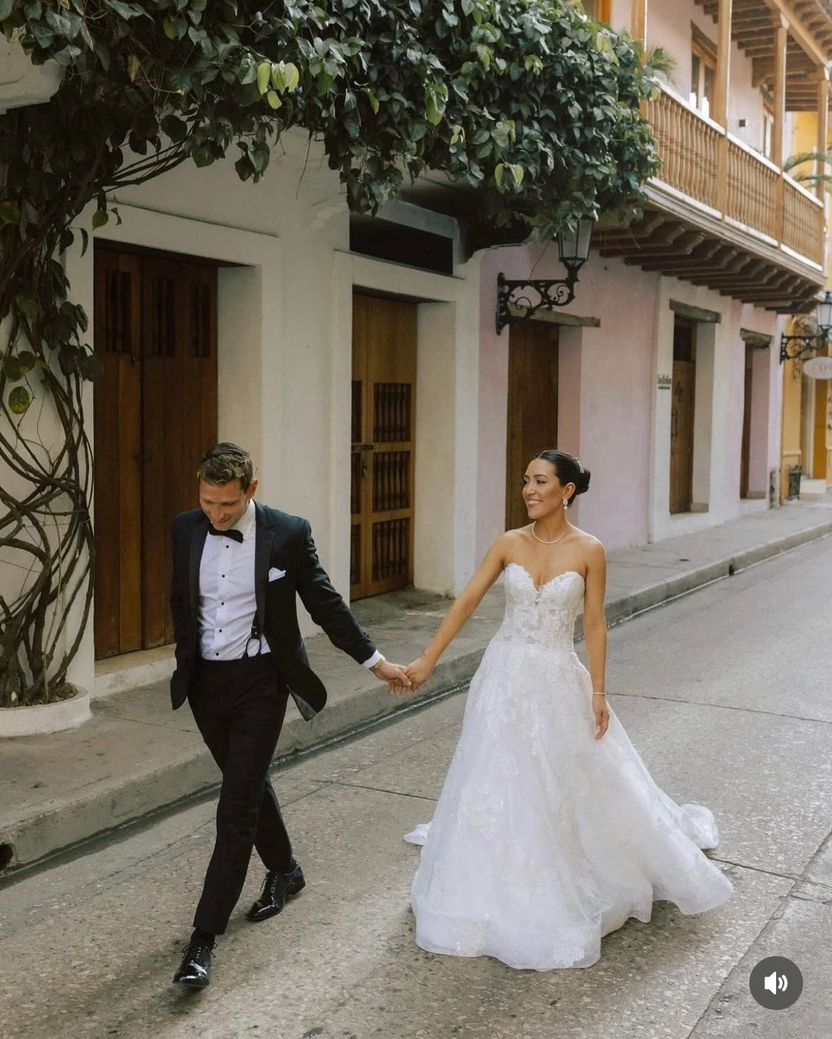 Bride and groom holding hands walking on a street with colorful buildings, bride in white wedding gown, groom in black tuxedo.