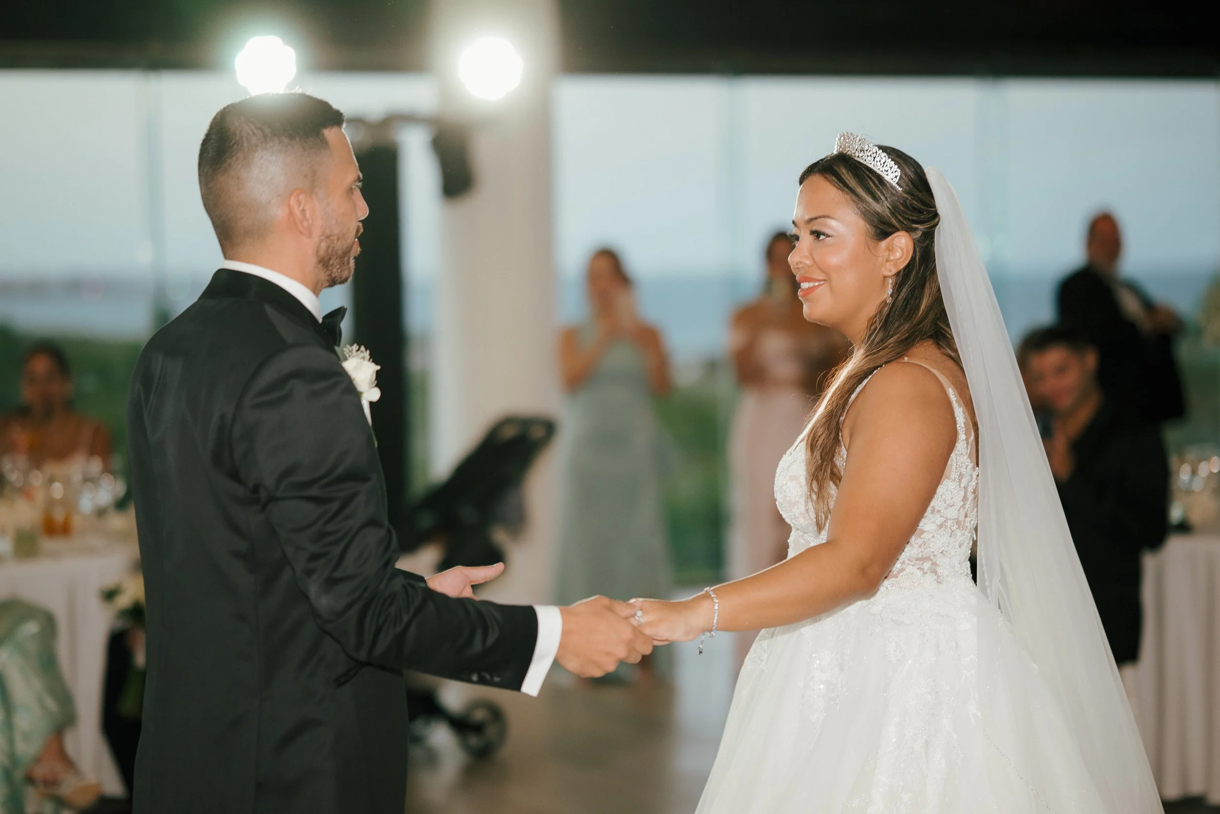 A bride and groom hold hands during their wedding ceremony. The bride is wearing a white lace wedding gown, a tiara, and a veil. The groom is dressed in a black tuxedo with a white shirt and bow tie. Guests can be seen in the background.