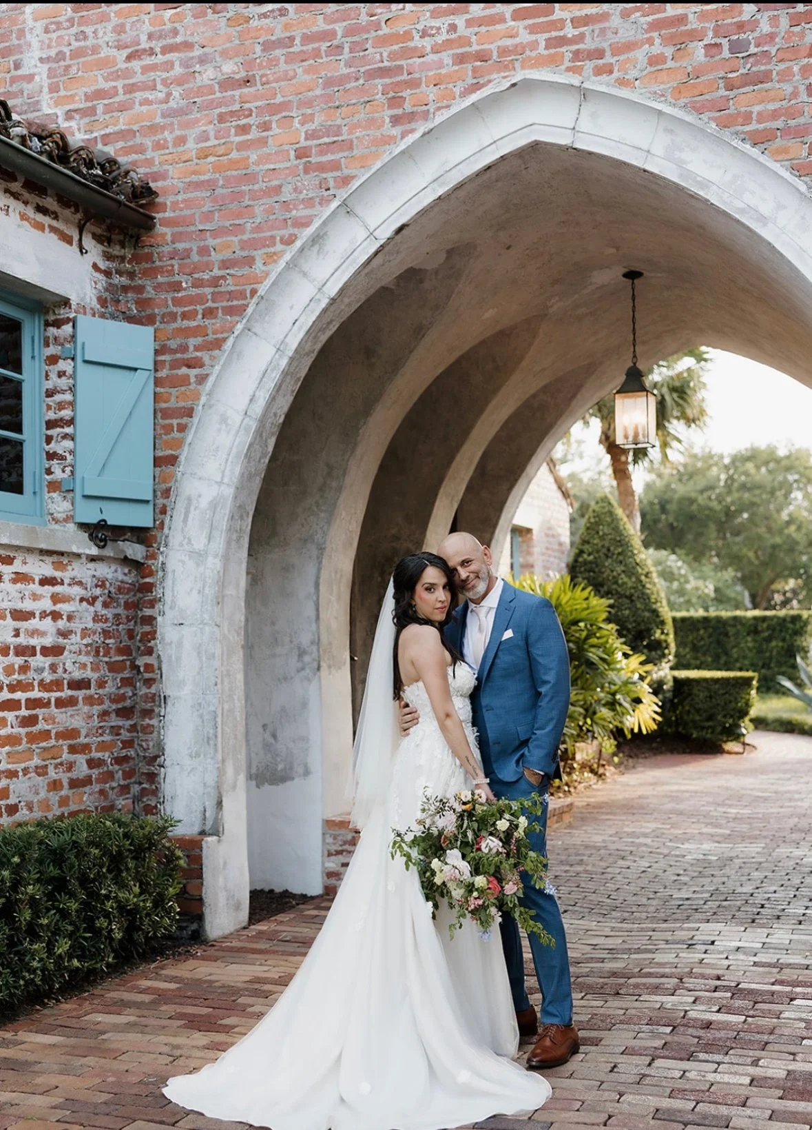 A bride and groom standing under an arched brick and concrete structure outdoors, with lush greenery in the background. The bride is wearing a white wedding dress and holding a bouquet, while the groom is in a blue suit and white shirt, smiling at the camera.