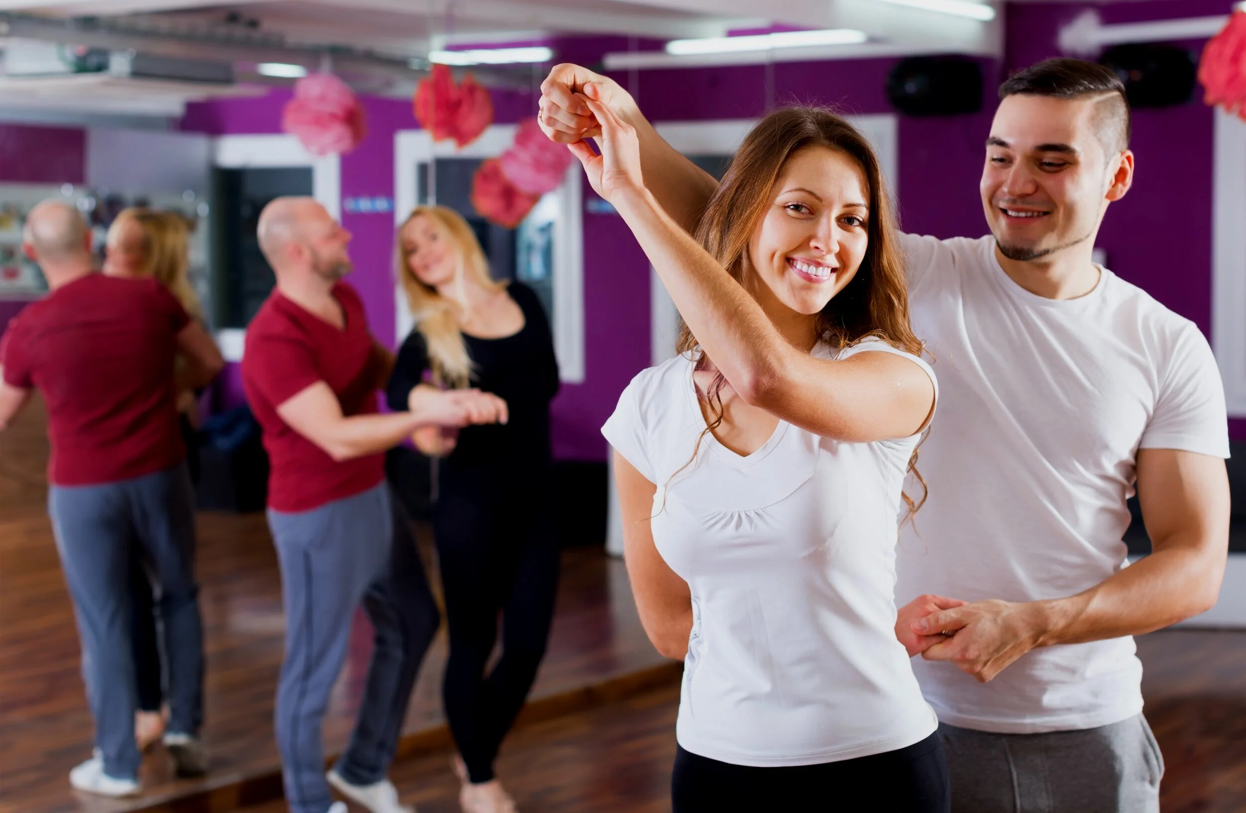 A young woman and man dancing together at a social event or dance class, with other couples dancing in the background, in a room decorated with pink paper lanterns and purple walls.