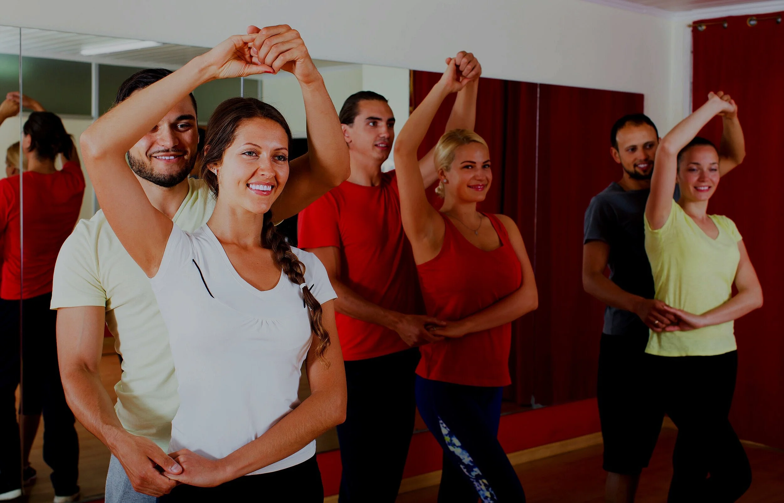 Group of six people in a dance class holding hands and smiling, with a mirror reflecting their image on the wall behind them.