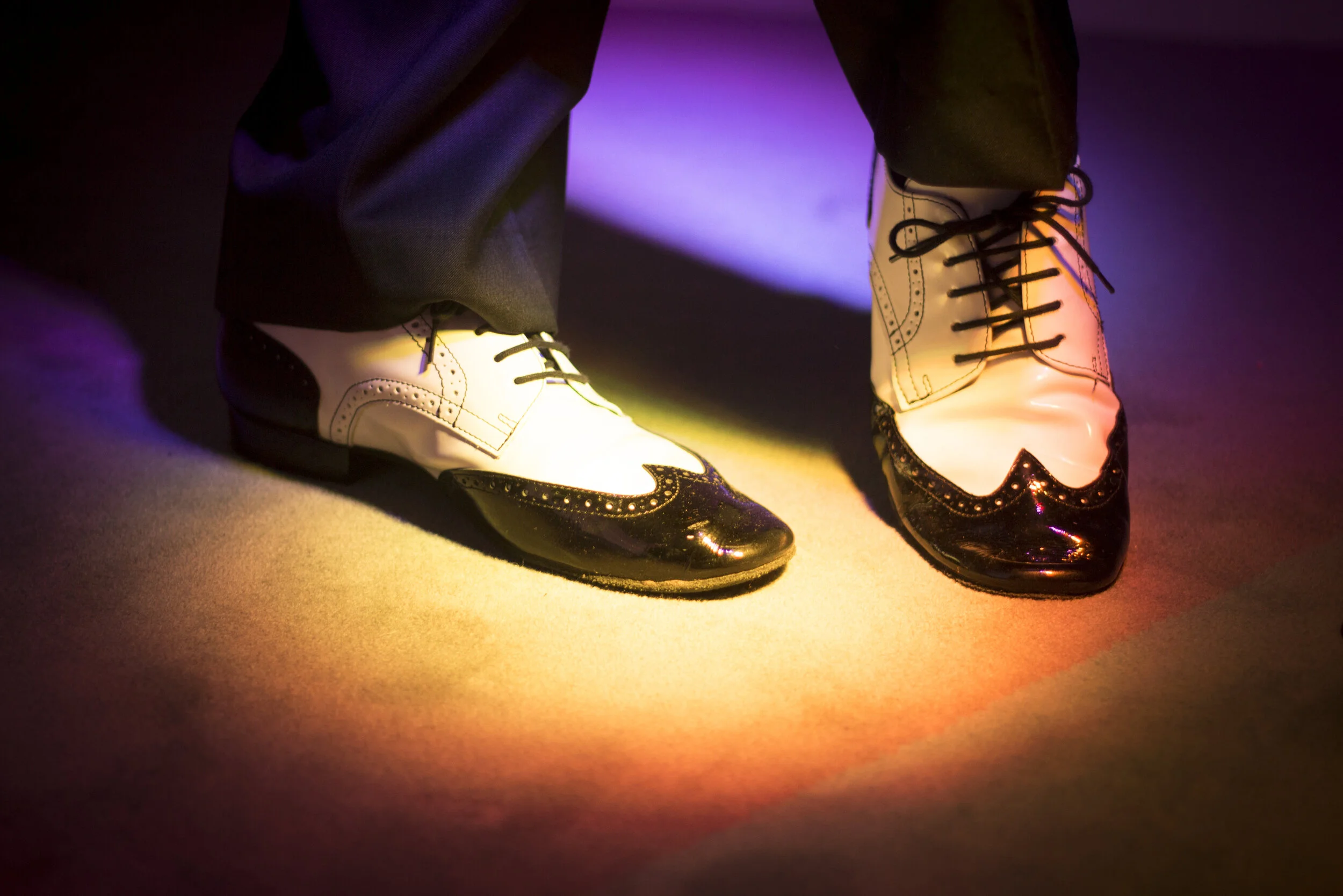 Close-up of someone's feet in black and white brogue shoes, standing on a colorful, illuminated floor.