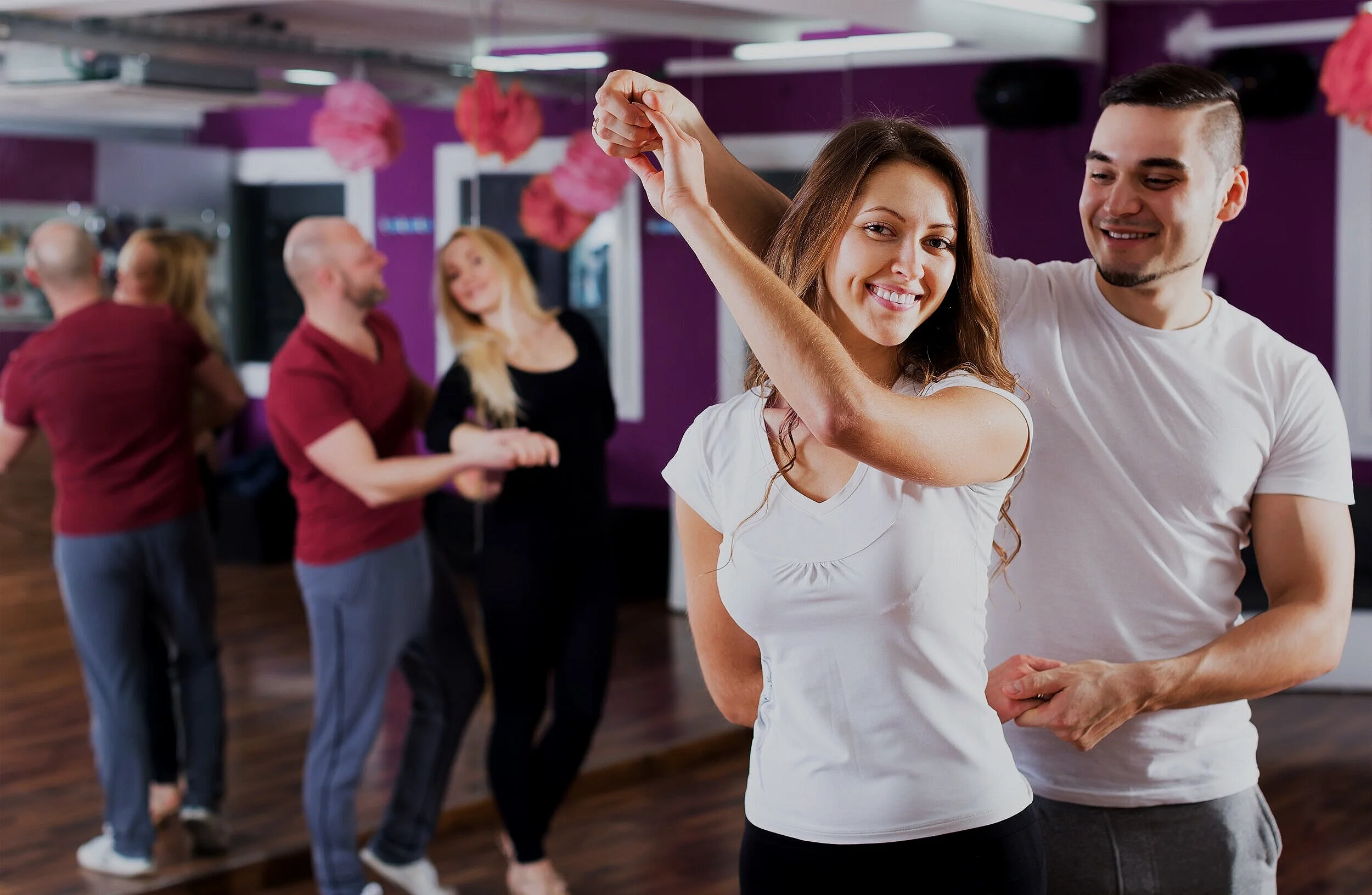 A couple dancing and smiling at a social gathering in a room decorated with pink paper lanterns, with other couples dancing in the background.