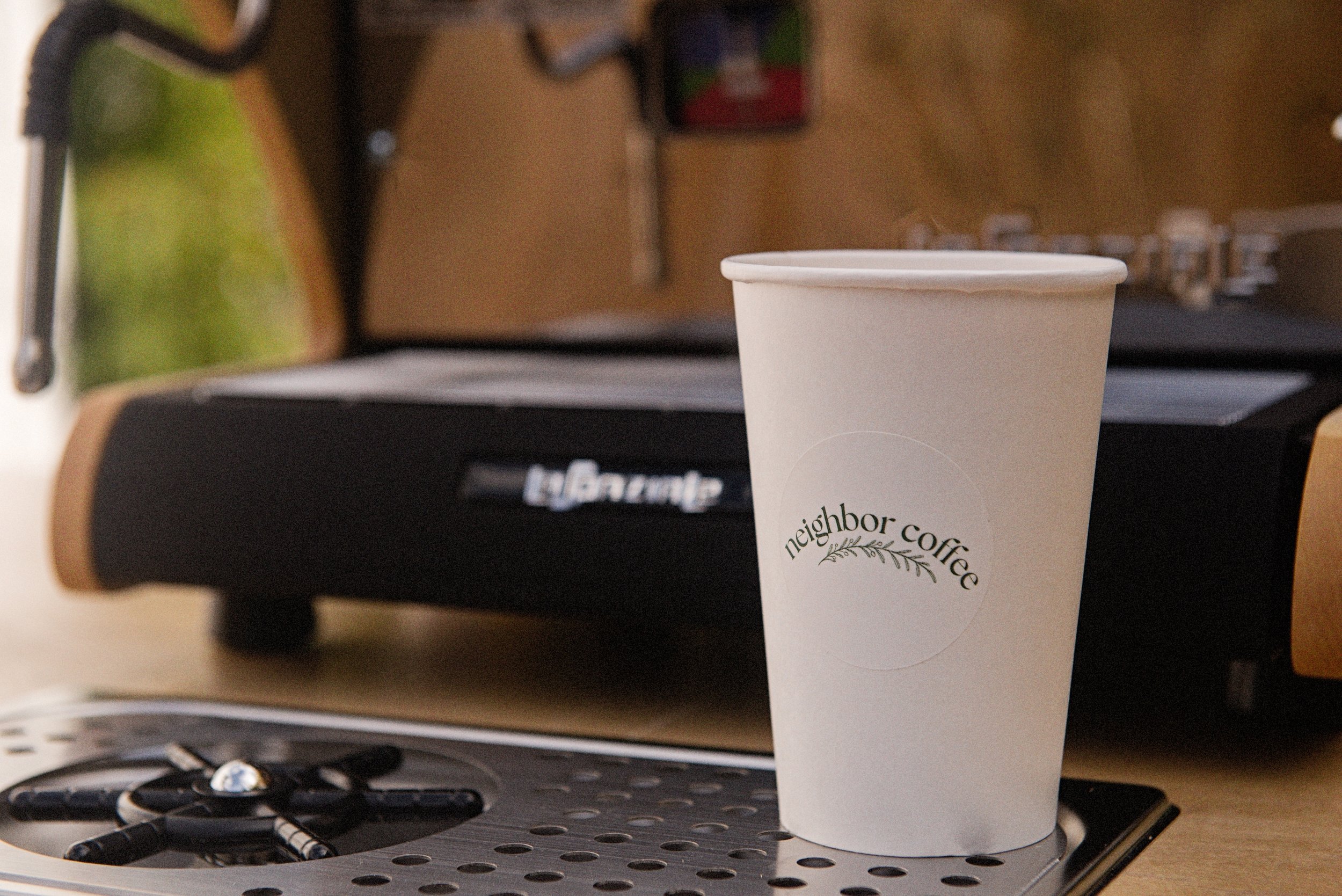 A white paper coffee cup with the logo "neighbor coffee" on it, placed on a coffee machine countertop.