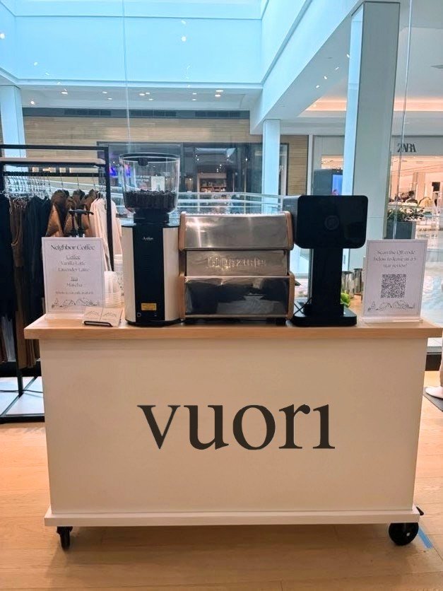 A coffee stand labeled 'vuori' inside a shopping mall, featuring a coffee grinder, a stainless steel coffee machine, and a digital order screen.
