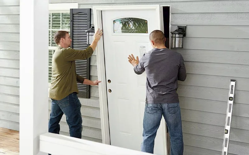Two men are installing a front door on a house. One man is holding the door with a window at the top, while the other is pushing it into place. A level tool is leaning against the wall.
