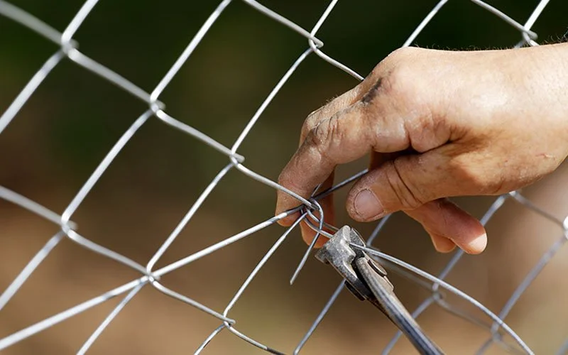 A person's hand using pliers to bend a wire on a chain-link fence.