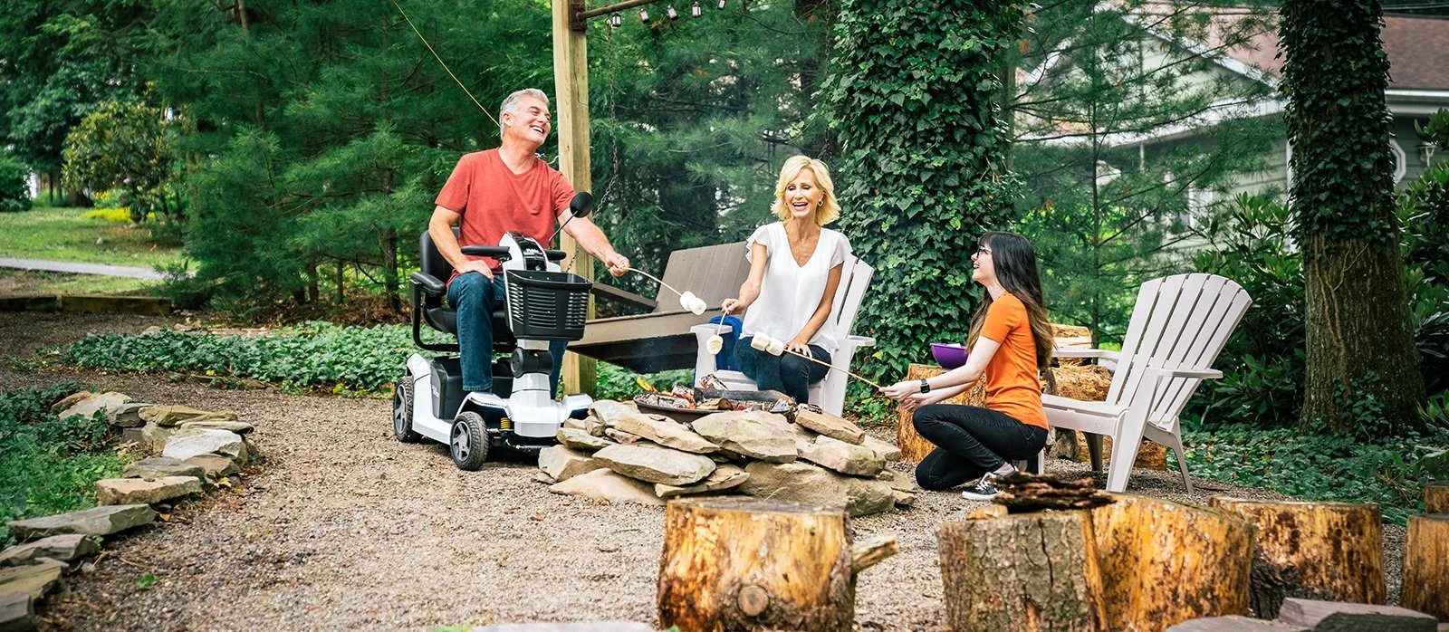 Three people, two women and one man, are outdoors around a small fire pit roasting marshmallows. The man is in a mobility scooter, and all three are smiling and enjoying the moment. The setting is a lush, green backyard with trees and seating.