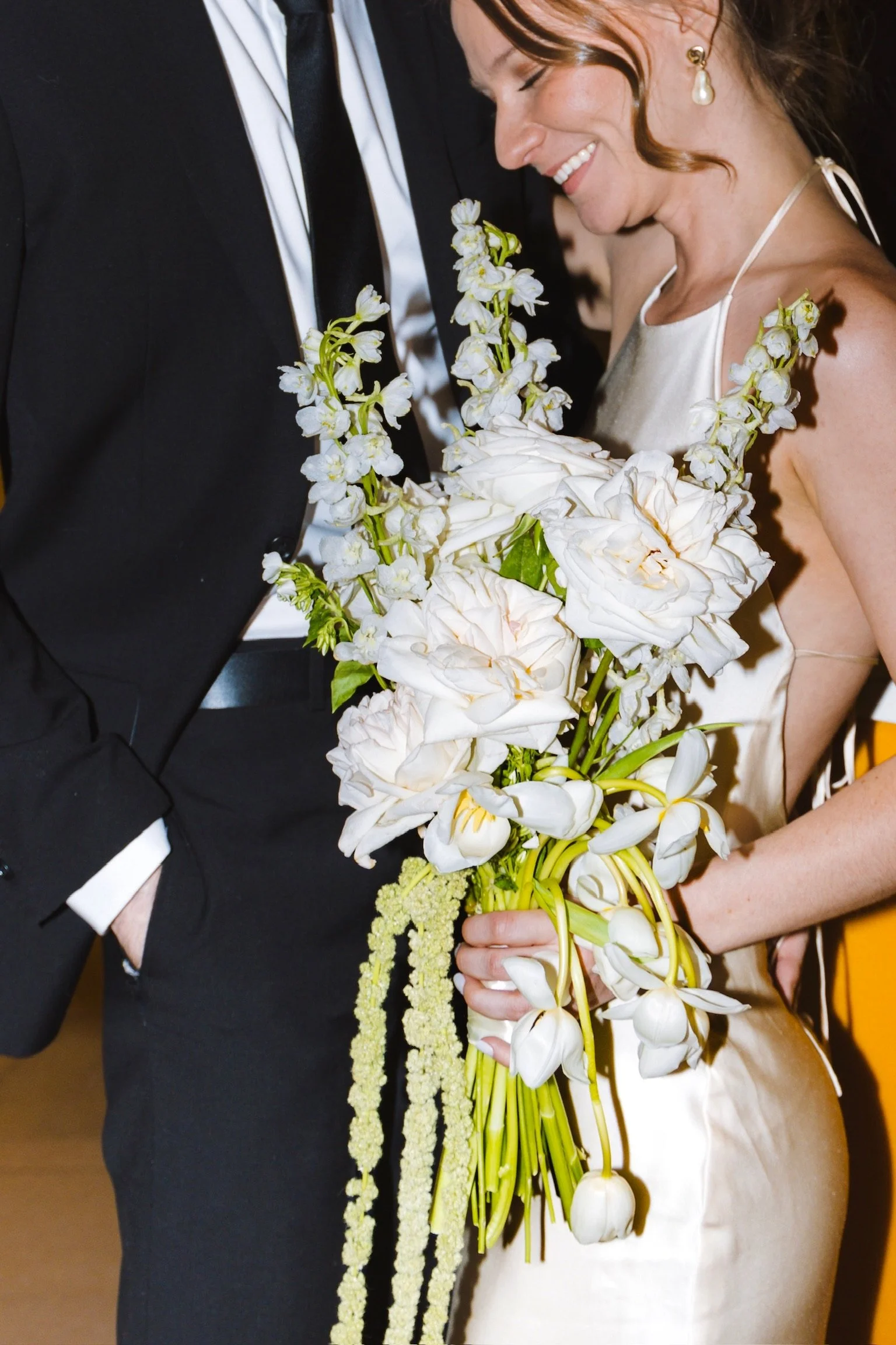 A bride and groom are holding a large bouquet of white flowers, smiling and embracing each other at a wedding celebration.