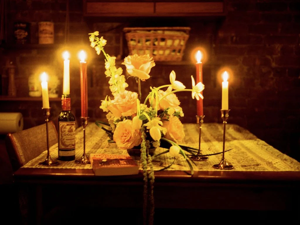 A dining table with a floral centerpiece, lit candles, and a book, set in a cozy, dimly lit room with a brick wall background.