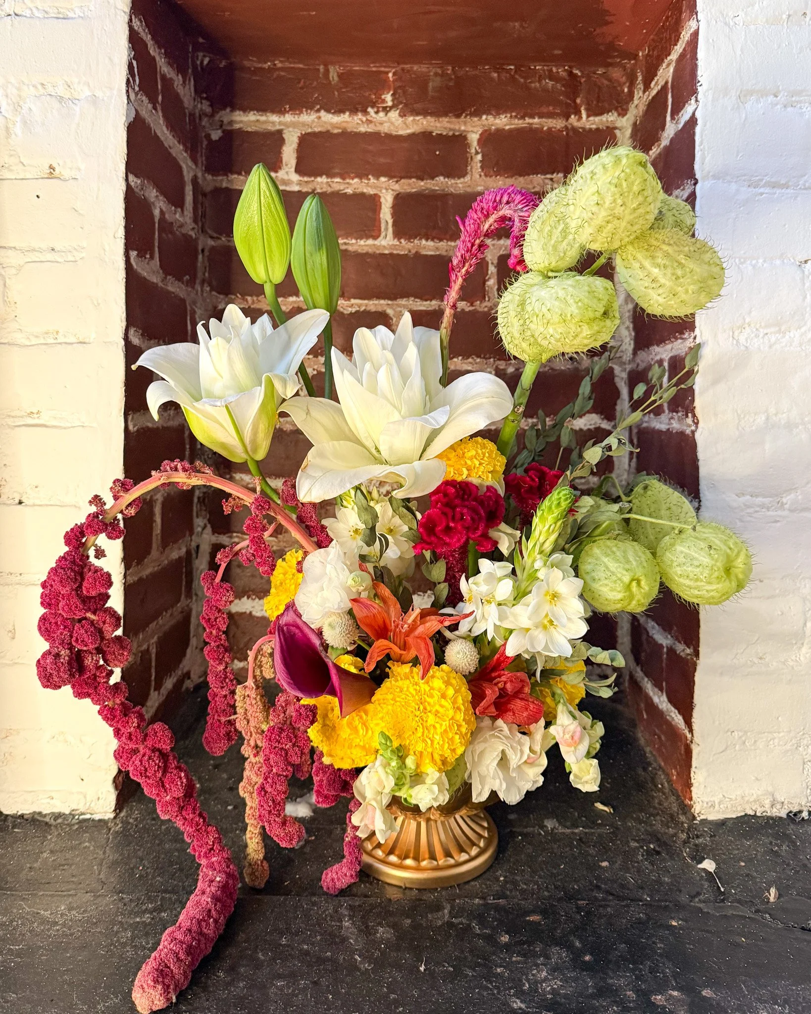 Colorful flower arrangement with white lilies, pink calla lilies, yellow marigolds, and other mixed flowers in a gold vase, placed on a dark surface near a brick fireplace.