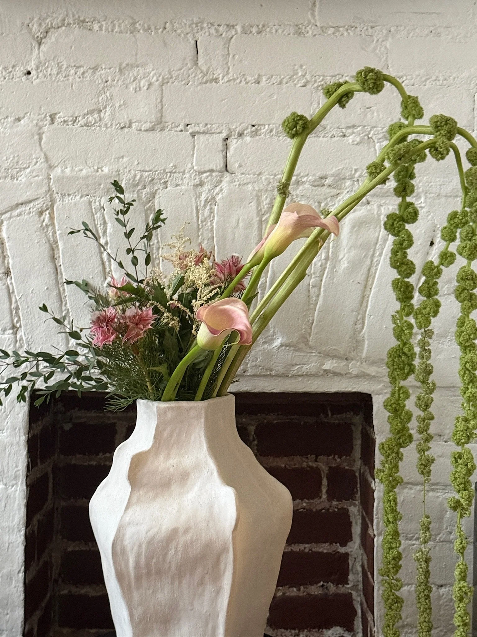A bouquet of flowers in a decorative beige vase against a white brick wall.