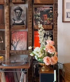 Flower arrangement with pink and white roses in a gold vase on a blue cabinet, bookshelf with vinyl records and framed photos in the background.