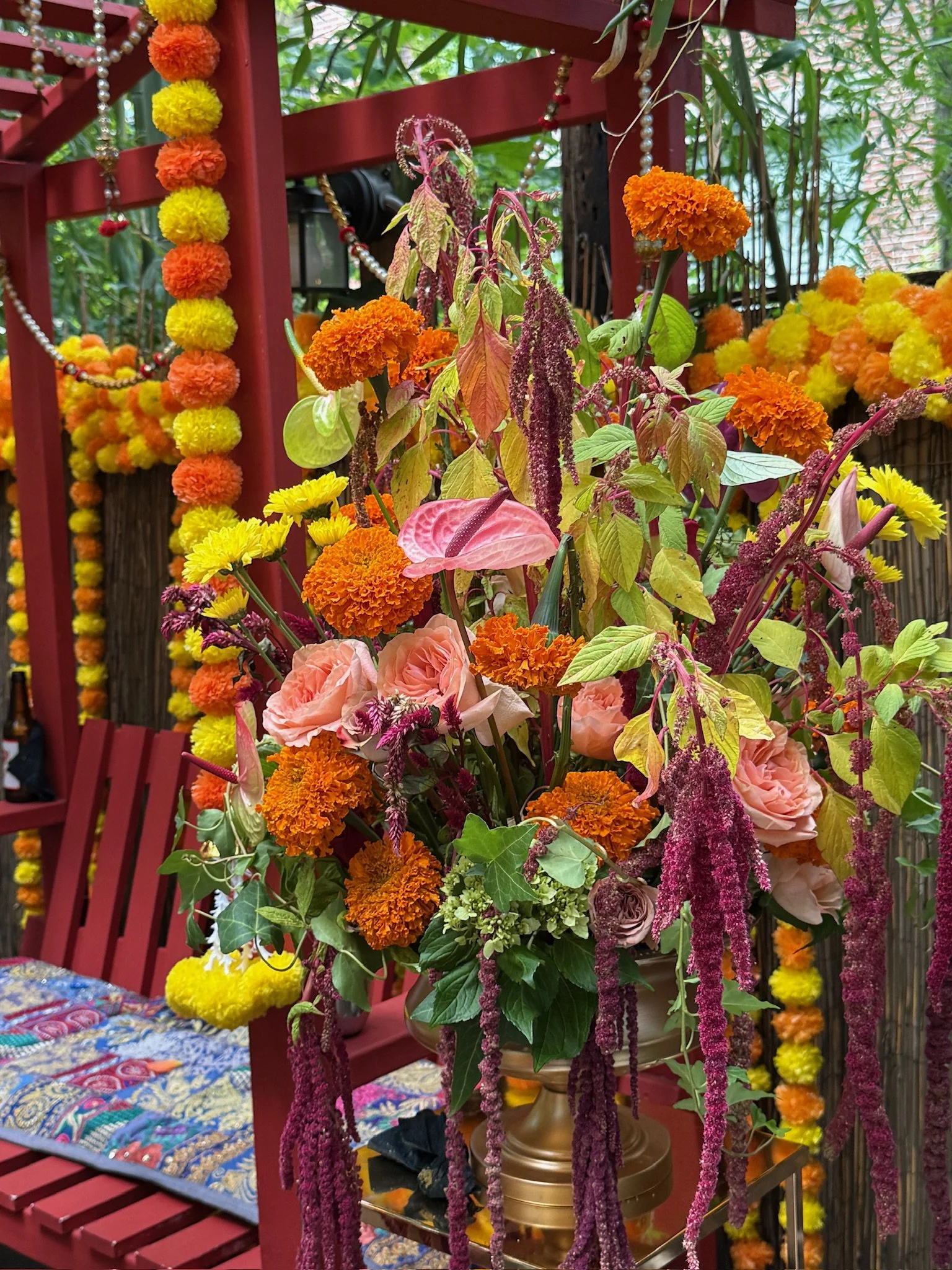 A floral arrangement with orange marigolds, yellow flowers, pink roses, purple amaranth, and green leaves displayed on a table with a red background decorated with marigold garlands.