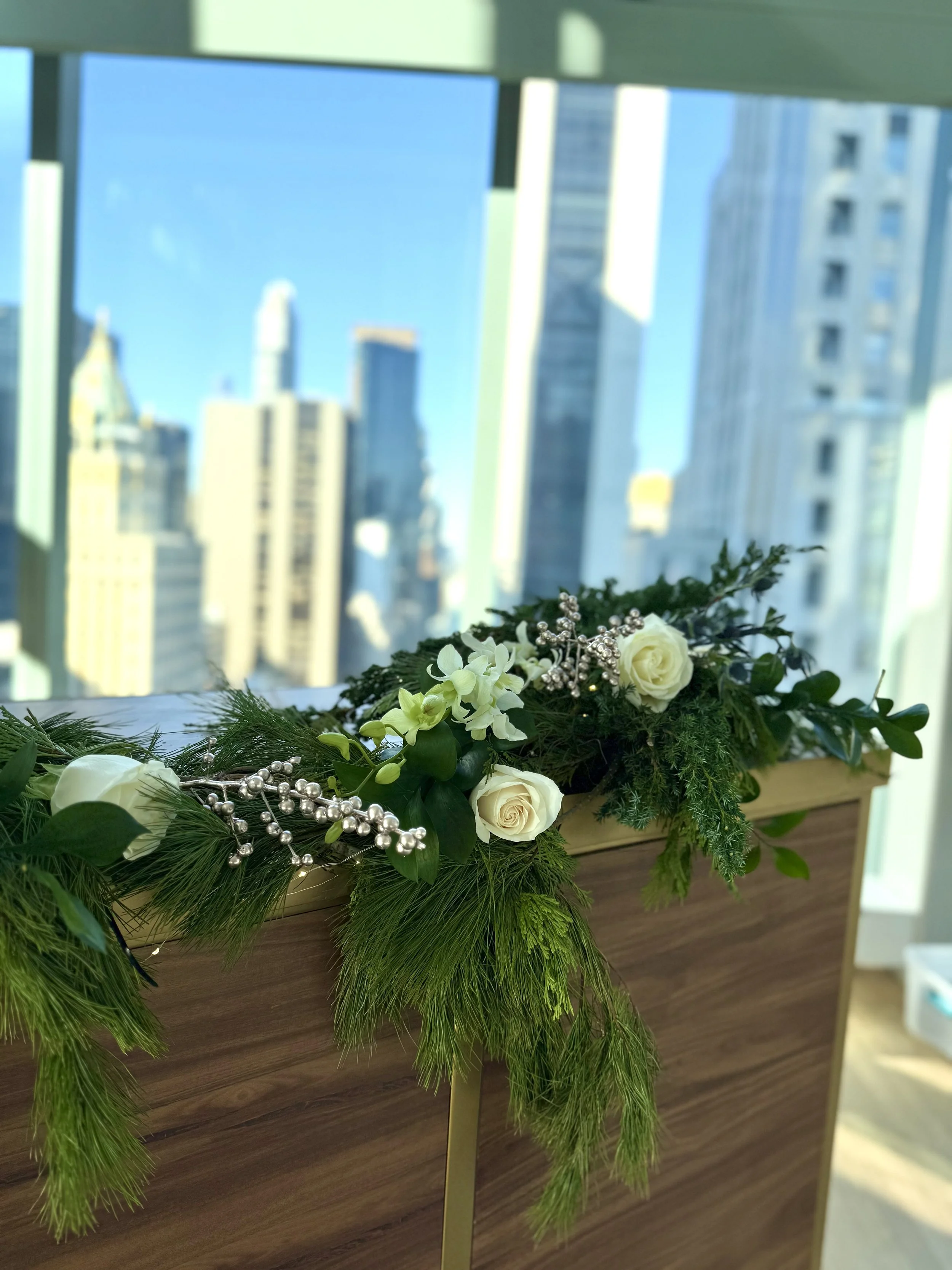 A floral arrangement with white roses, green foliage, and silver decorative beads on a wooden surface with a city skyline view in the background.