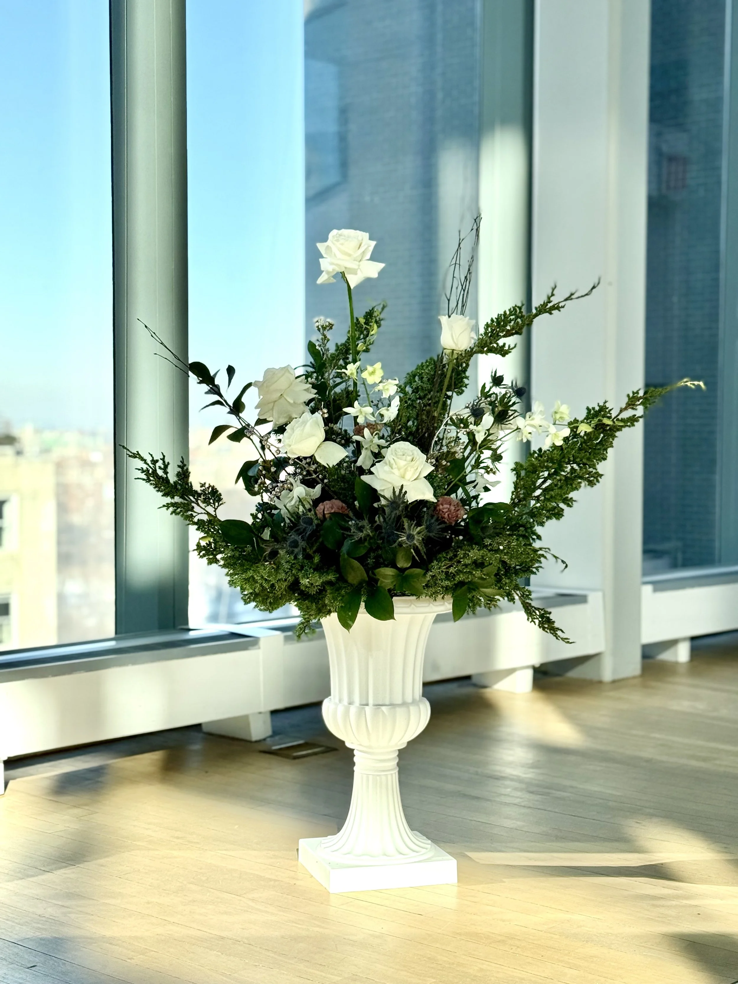 A white decorative flower vase filled with white roses, assorted greenery, and small flowers, placed on a wooden floor near large windows with a cityscape view.