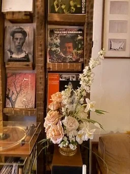 Decorative arrangement of white, pink, and beige flowers in a glass vase on a table, with a bookshelf filled with vinyl records and books in the background.