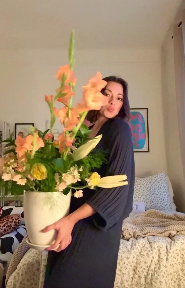 A woman with black hair poses for a photo holding a large white vase of flowers, including white lilies and orange gladiolus, in a bedroom.
