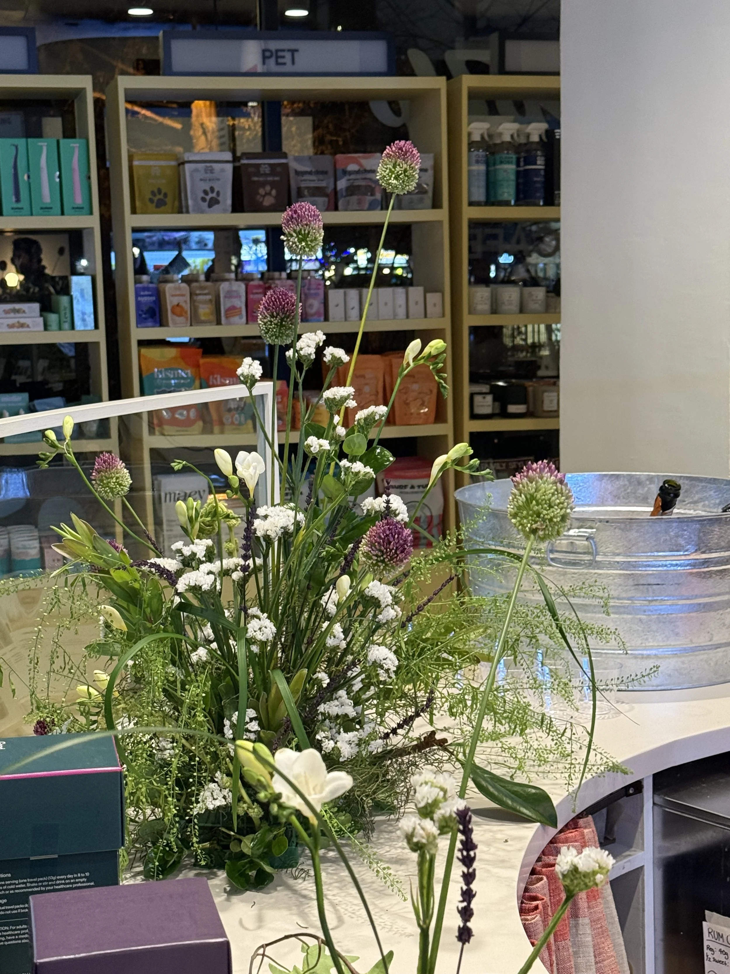A floral arrangement with white, purple, and green flowers on a counter in a store, with shelves of pet products in the background.