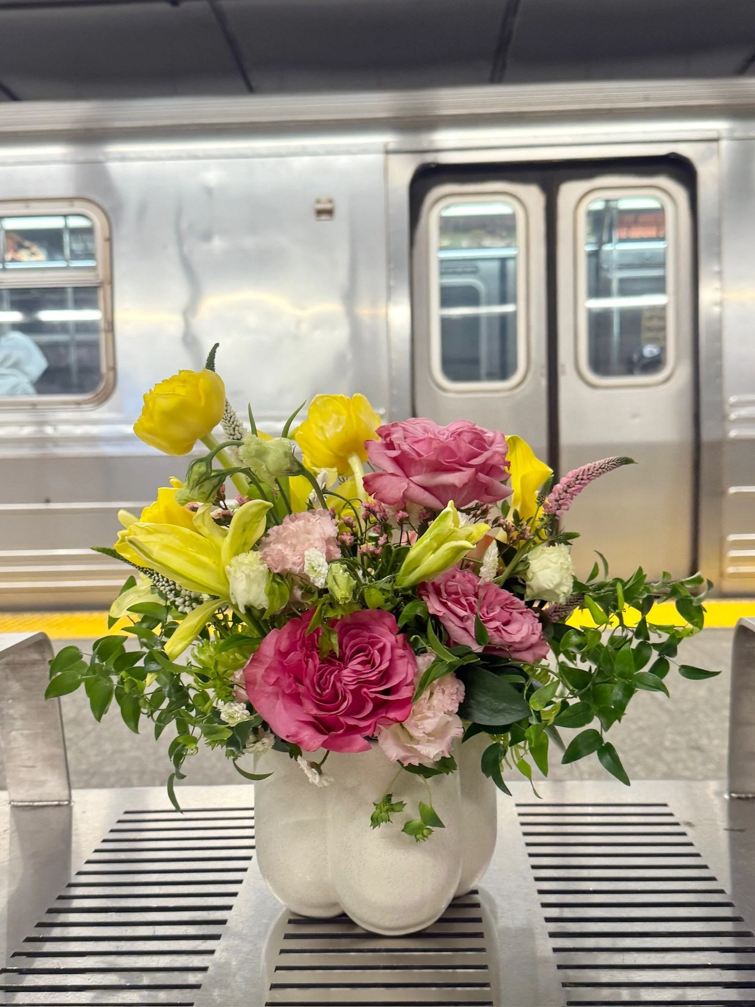 A bouquet of pink, yellow, and white flowers in a white vase, placed on a metallic surface, with a metal train door and windows in the background.