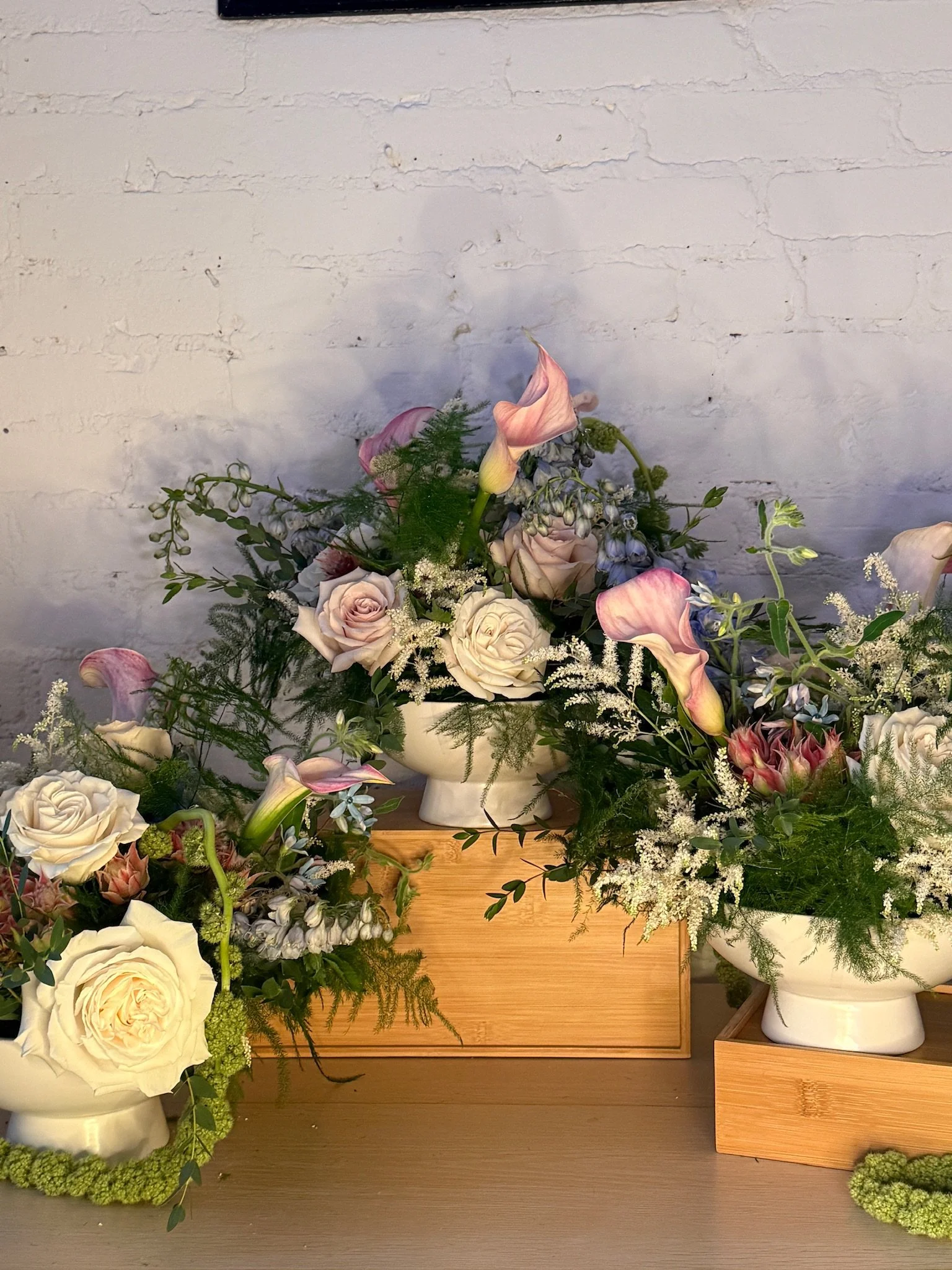 A floral arrangement with roses, calla lilies, and other flowers in white vases situated on wooden blocks against a white brick wall background.