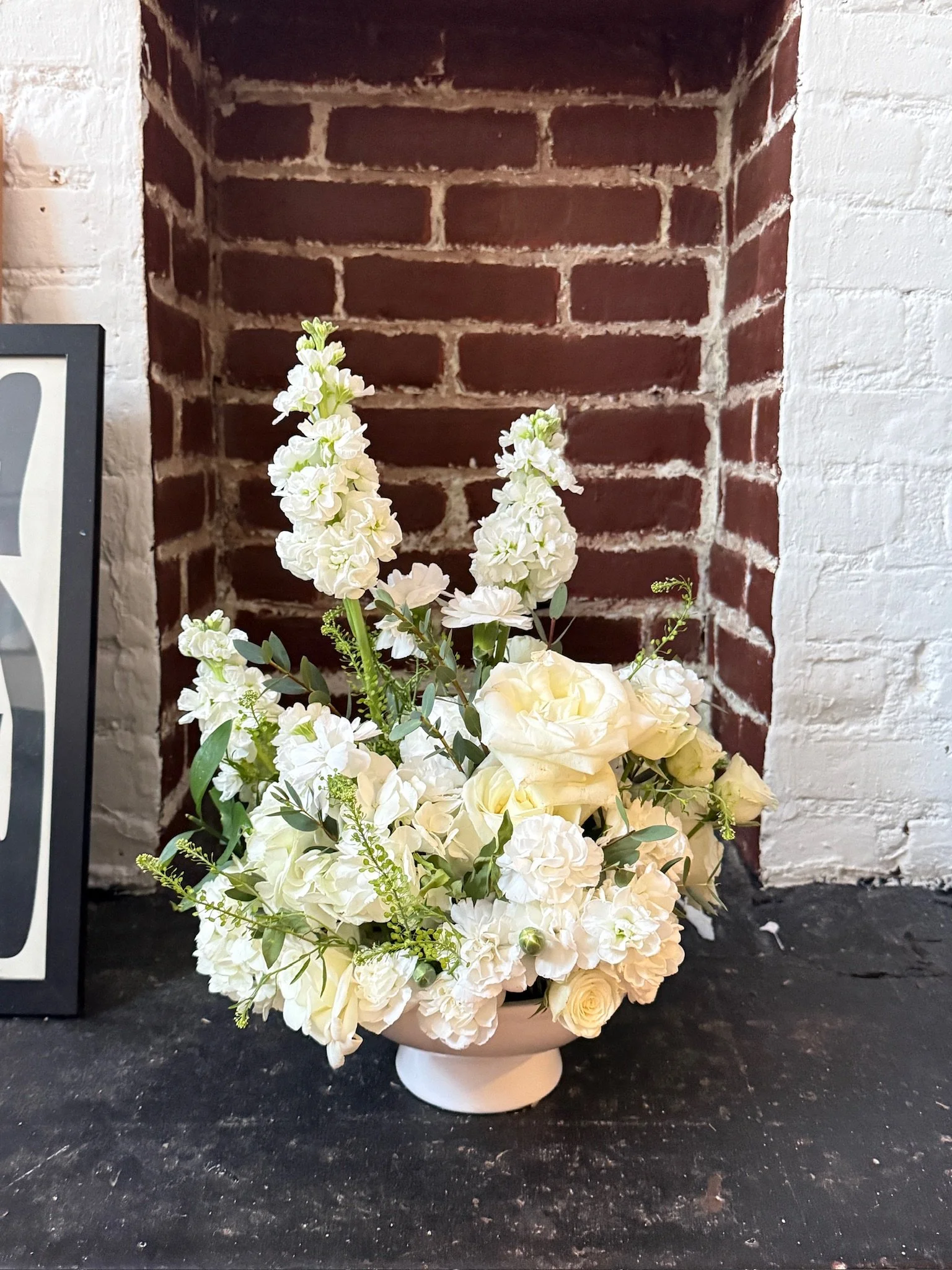 A white flower arrangement with roses and other flowers in a white vase on a black surface, with a brick wall background.