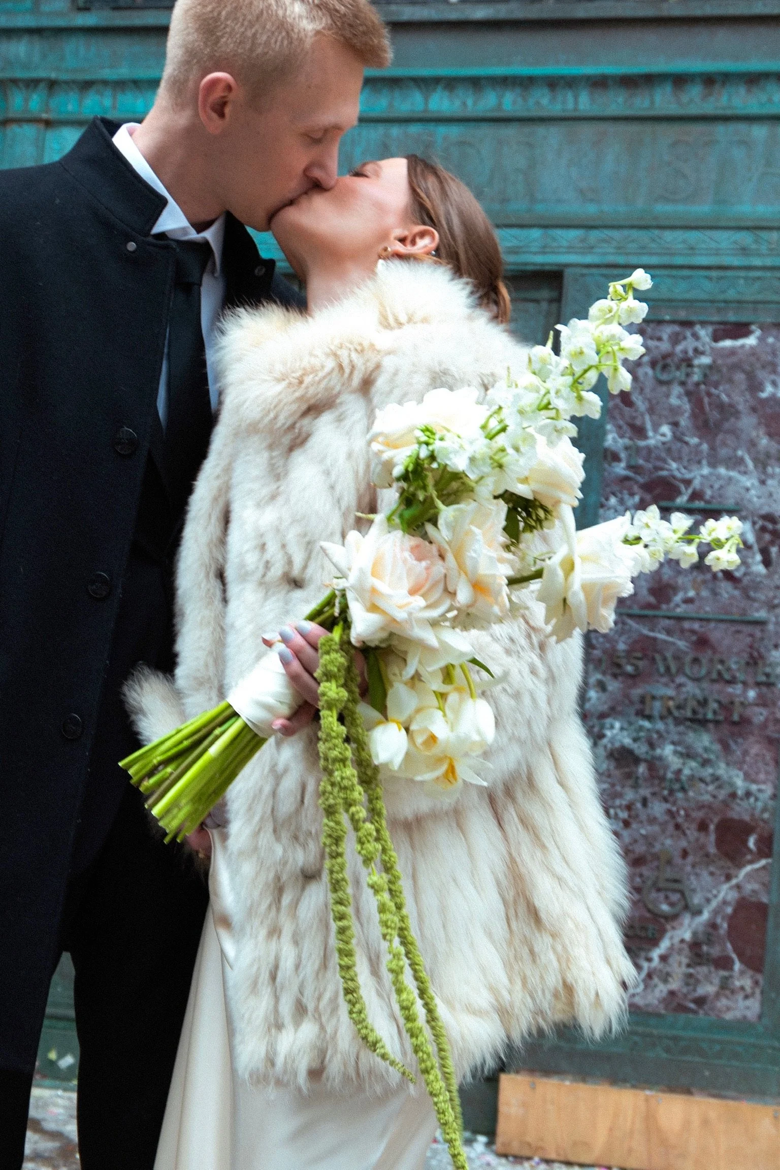 A couple kissing, with the woman holding a bouquet of white and cream flowers, standing outdoors in front of a weathered wooden wall.