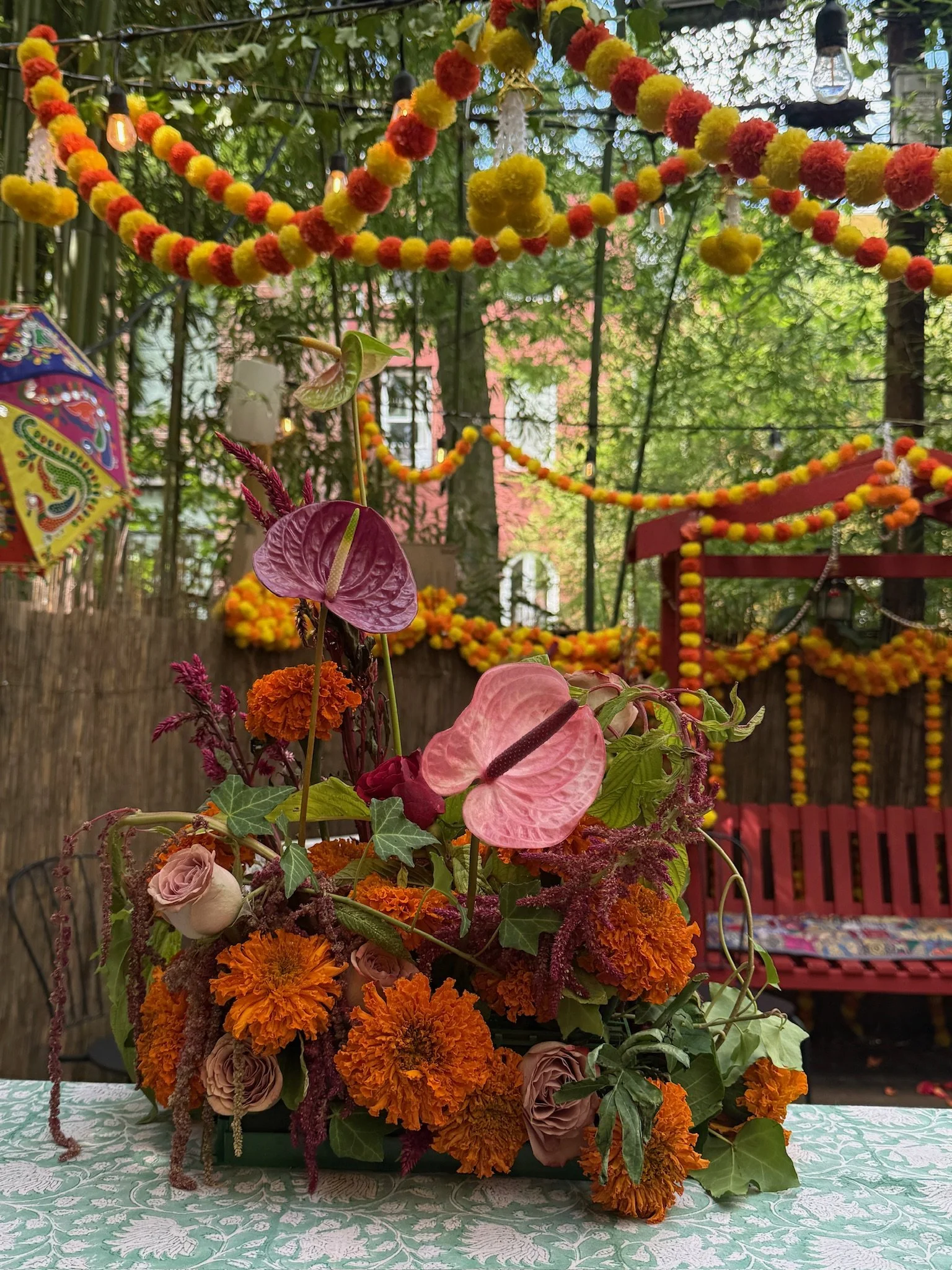 Decorative floral arrangement on a table with orange marigolds, pink anthuriums, roses, and greenery, set outdoors with colorful hanging garlands and a red bench in the background.