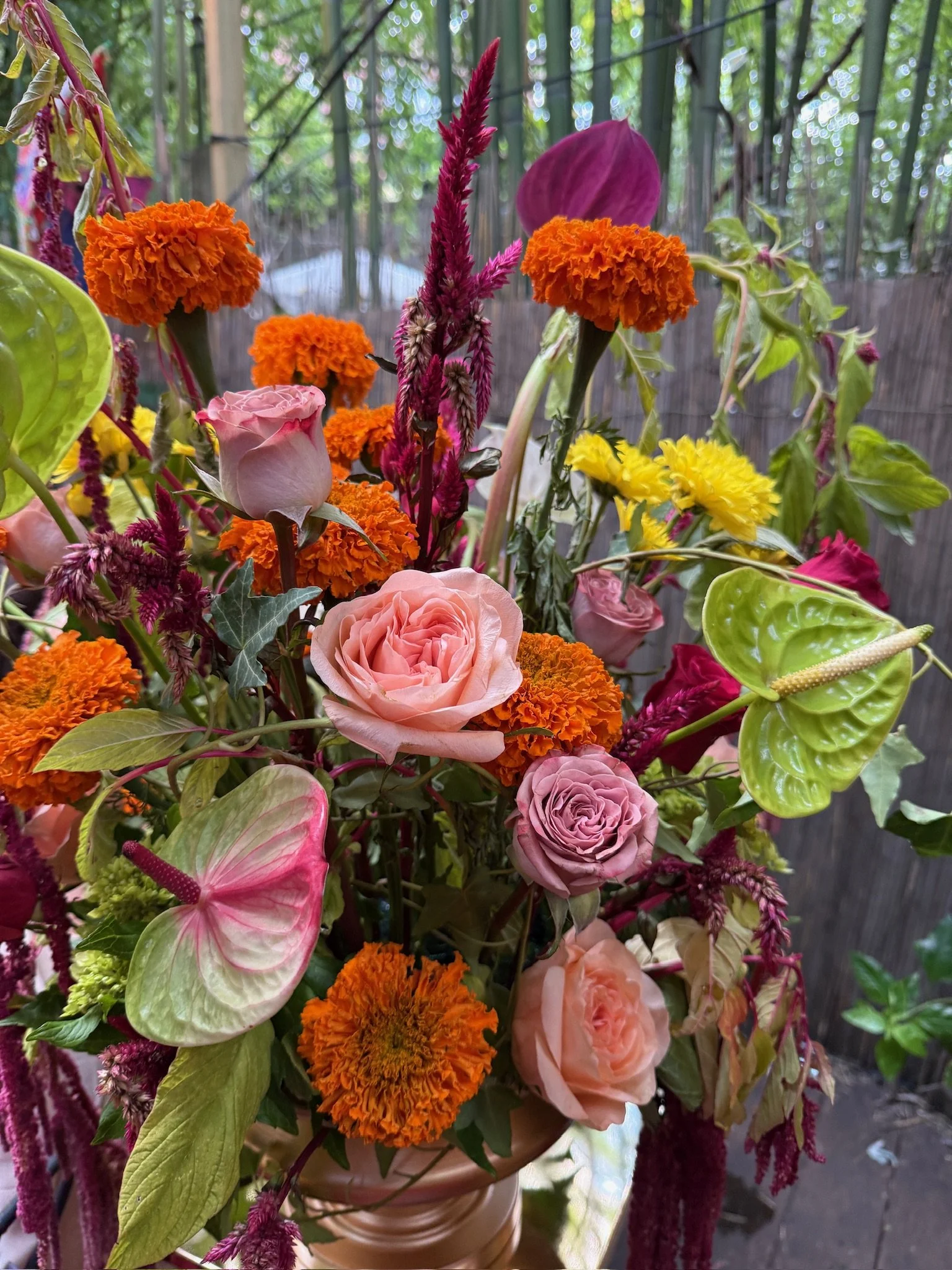 Close-up of a vibrant flower arrangement with pink roses, orange marigolds, yellow daisies, purple and pink anthuriums, accented with green leaves