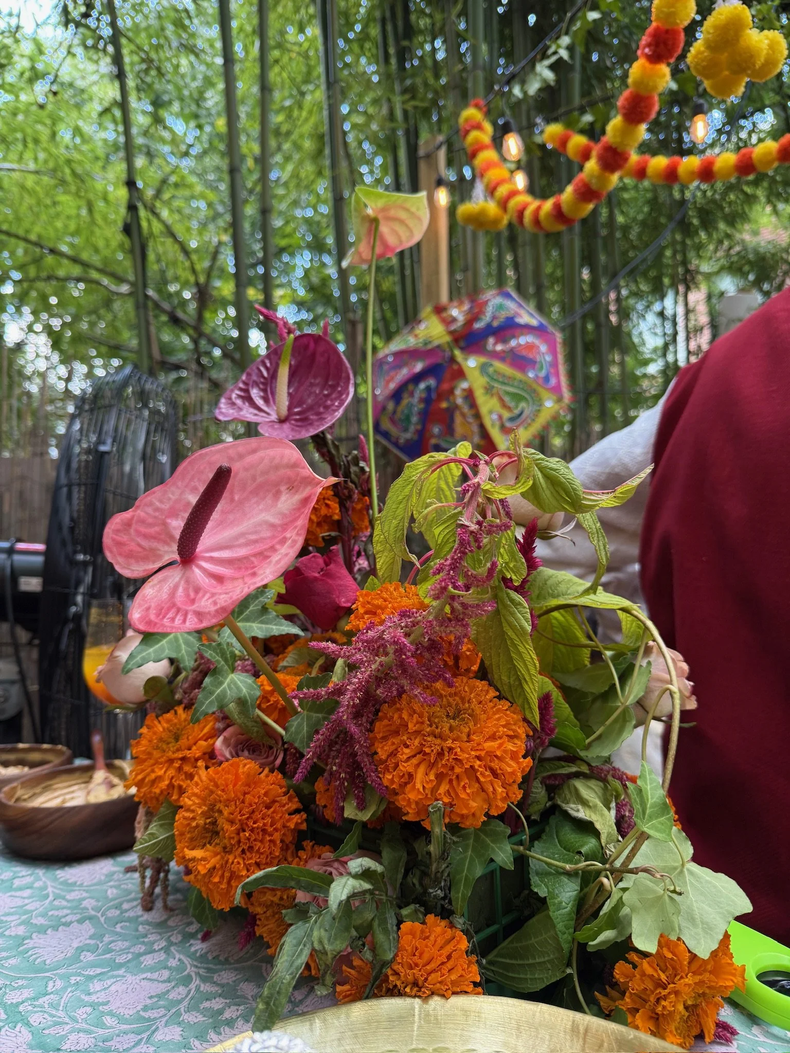 Colorful flower arrangement with marigolds, pink anthuriums, and green leaves, decorated with yellow and red pom-pom garland and string lights, against a background of green trees.