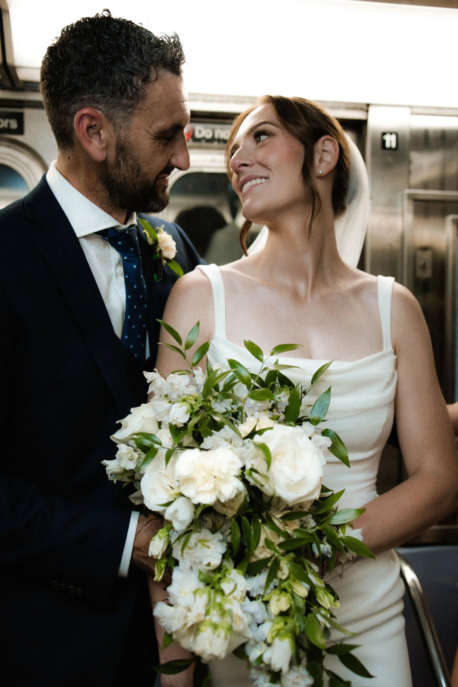 A man and a woman in wedding attire are looking into each other's eyes and smiling, with the woman holding a bouquet of white flowers and greenery.