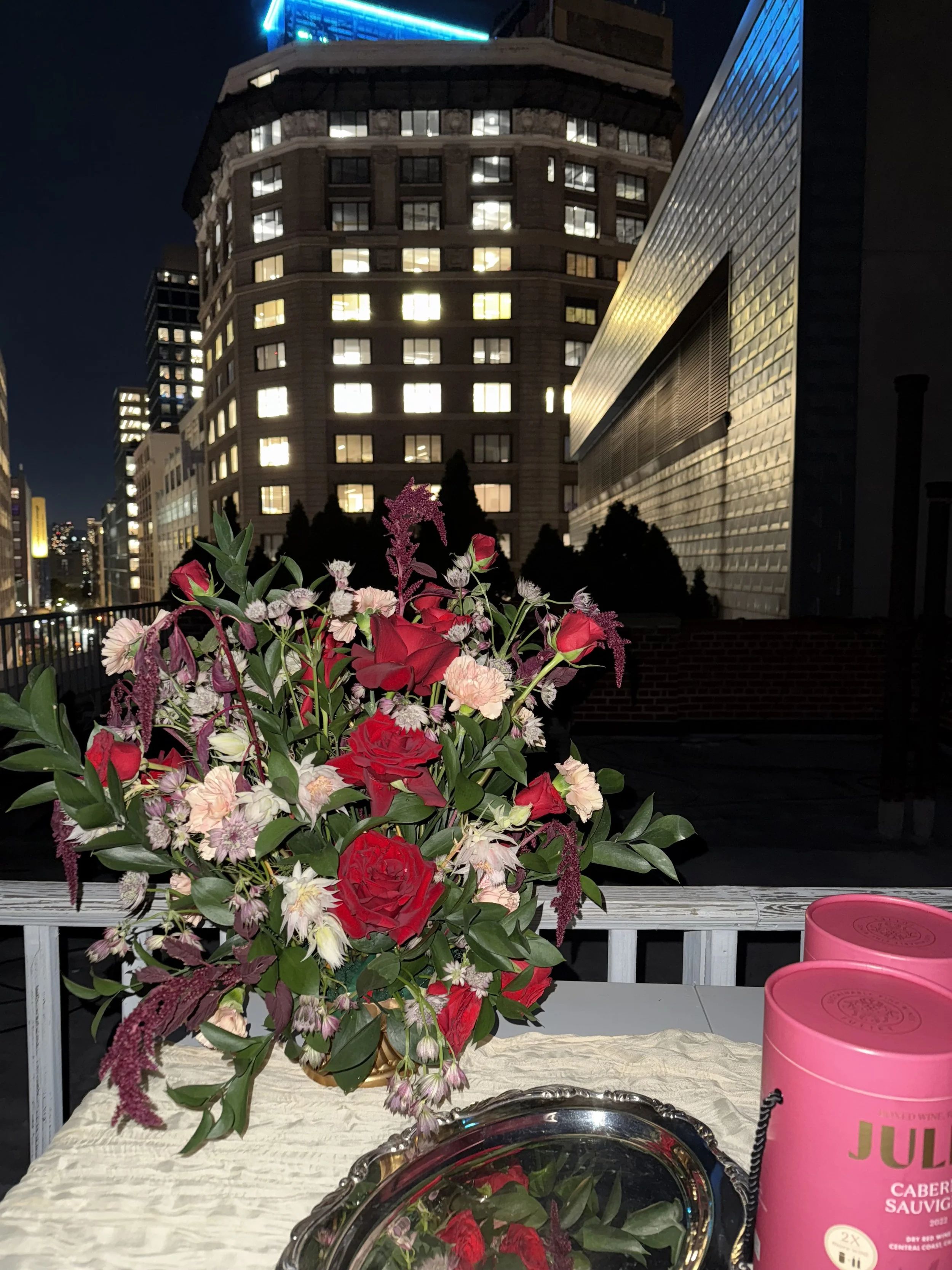 A bouquet of red, pink, and white flowers on a white table, with pink cans and a silver tray, against a cityscape at night with illuminated buildings.