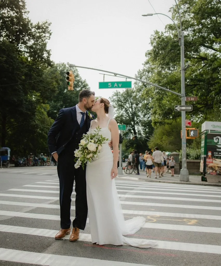 A bride and groom sharing a kiss on a crosswalk in an urban setting, with trees and street signs overhead.