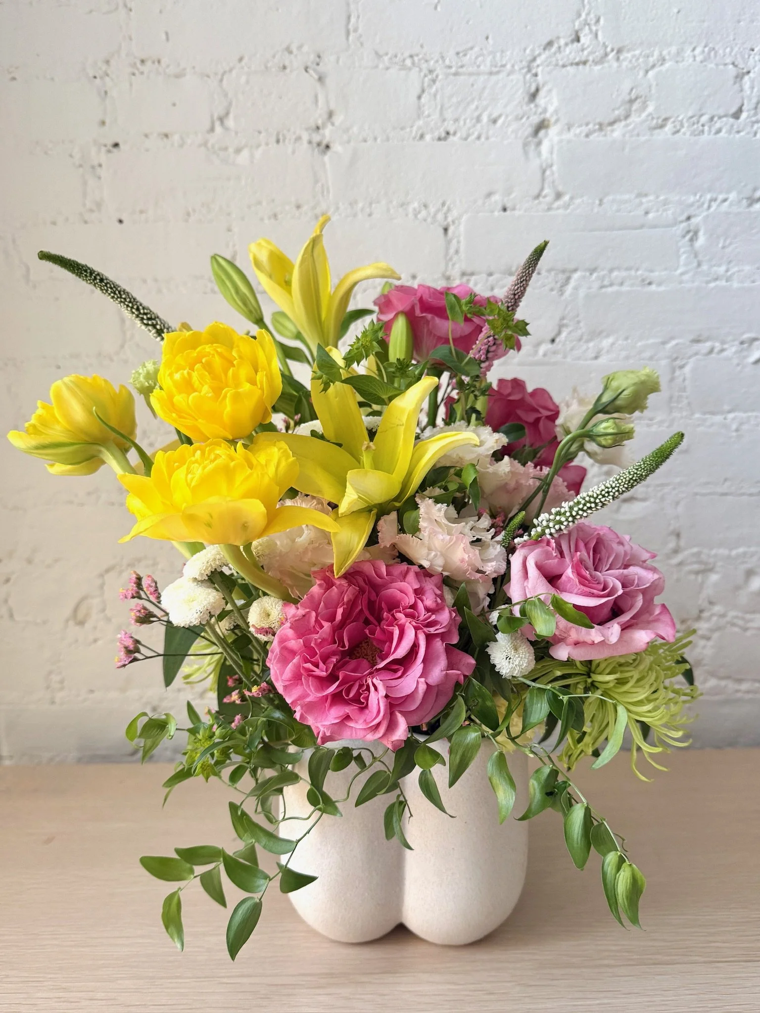 A white vase filled with a colorful arrangement of pink, yellow, and white flowers, with green foliage, placed on a light-colored wooden surface against a white brick wall background.