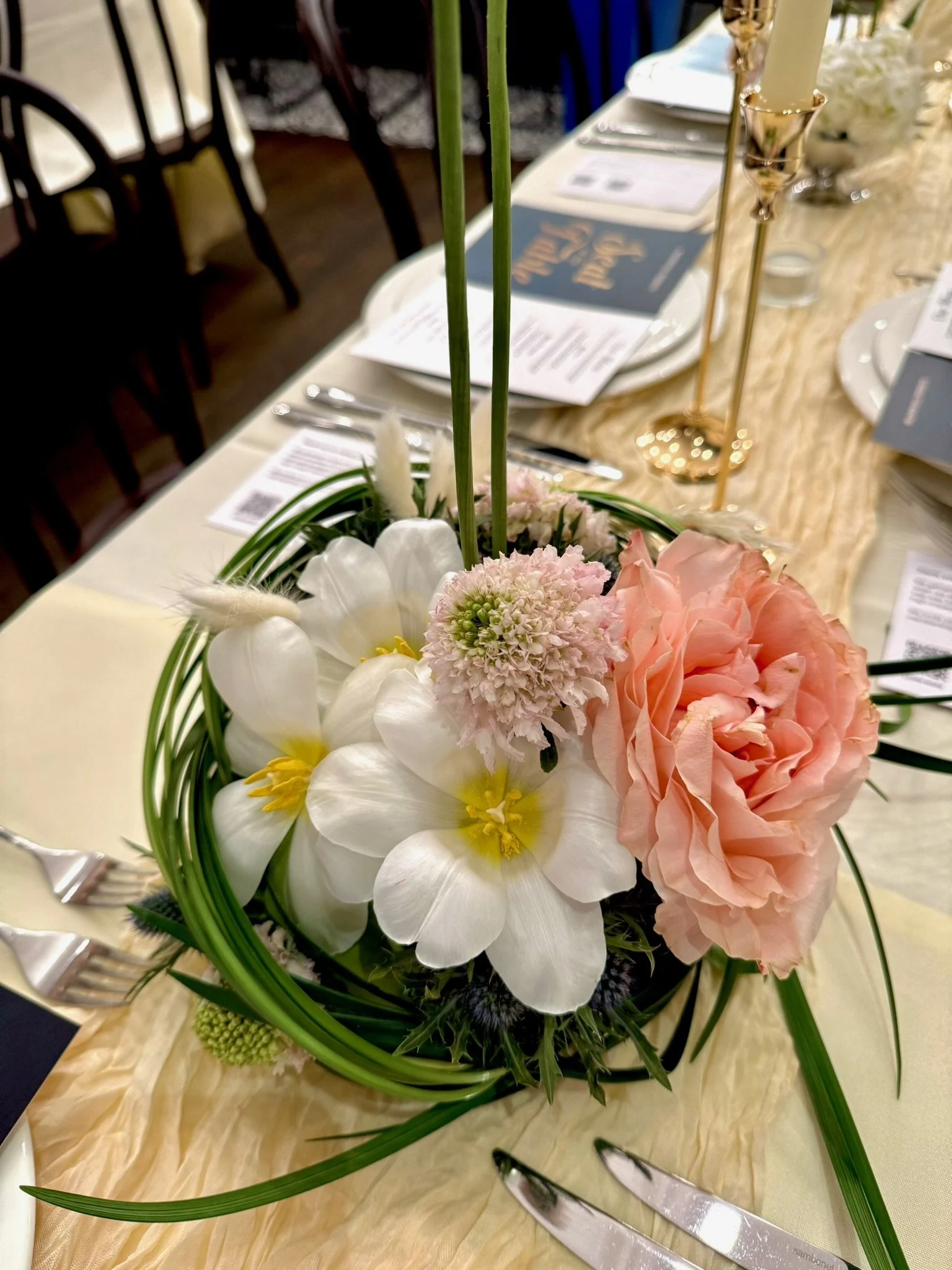 Elegant floral centerpiece with white orchids, pink carnations, and greenery on a table with gold accents and table settings.