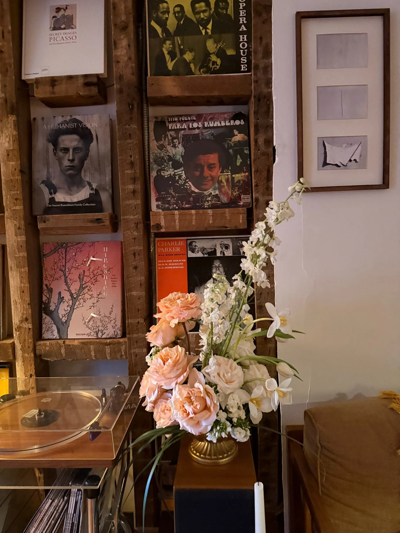 A large floral arrangement with white orchids and peach roses in a gold vase on a wooden table in a room decorated with vintage posters and artwork, including a black-and-white portrait of a young woman, black and yellow photographs of jazz musicians