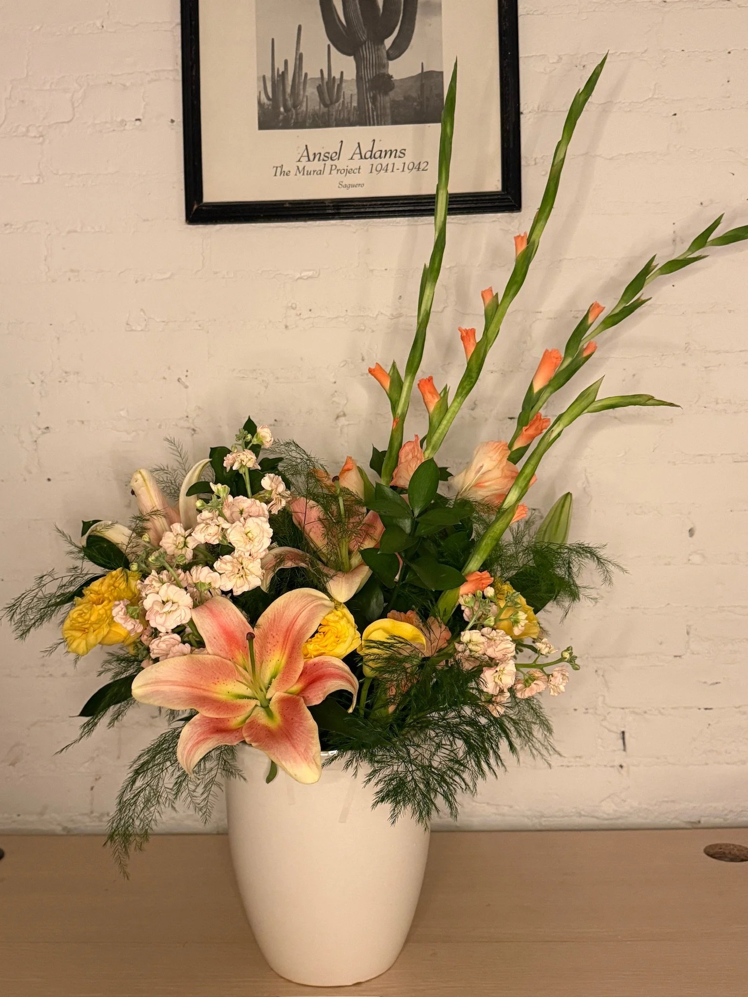 A floral arrangement in a white vase features pink lilies, peach gladiolus, white and yellow flowers, and green foliage, set against a white brick wall with a framed black-and-white picture of a cactus.