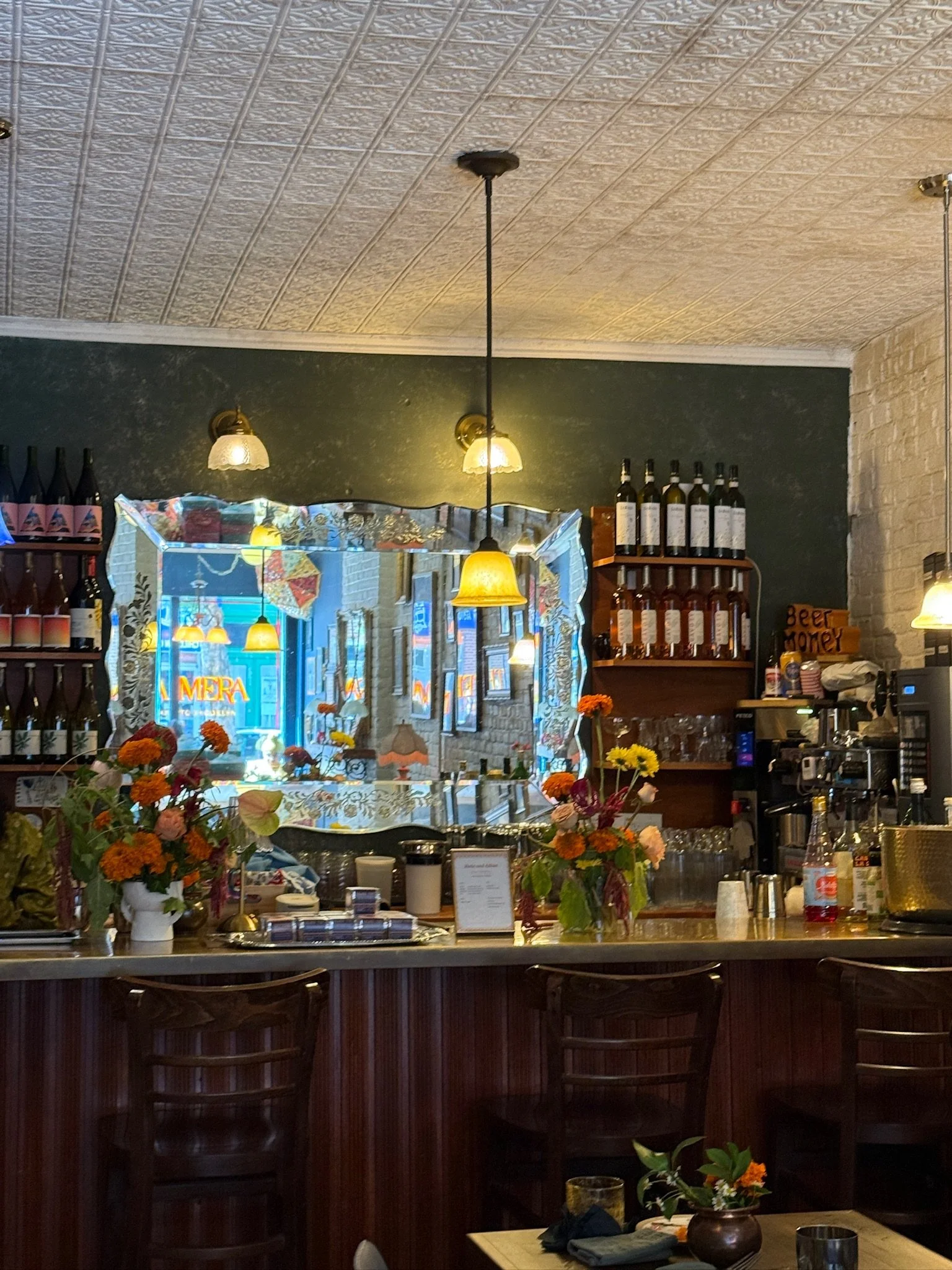 Interior view of a cozy cafe or restaurant with a bar counter, flower arrangements on the counter, bottles of wine and other beverages, hanging pendant lights, framed pictures on the wall, and a window with floral curtains and a neon sign outside.