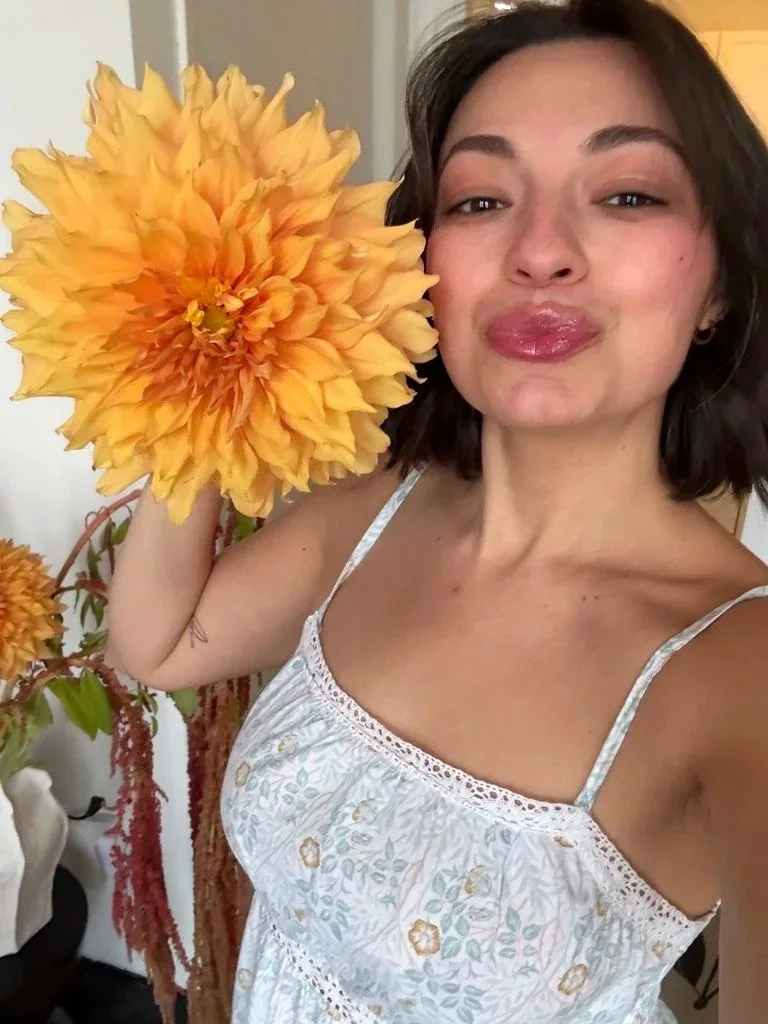 A young woman with shoulder-length dark hair holding a large yellow flower close to her face, wearing a white dress with floral embroidery.