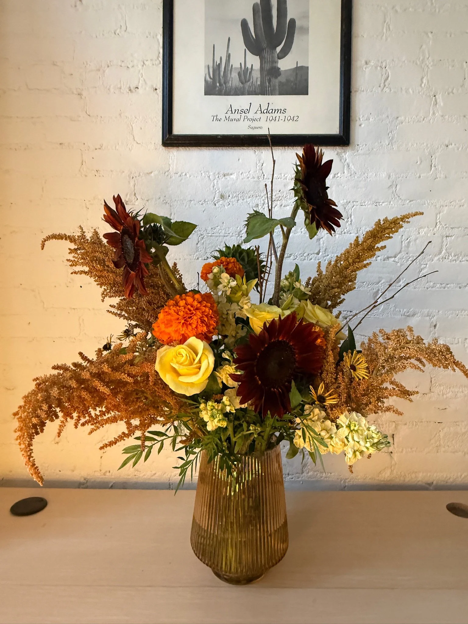 A tall glass vase with vertical ridges contains a large arrangement of flowers, including yellow roses, orange marigolds, purple and red sunflowers, and various greens, placed on a white table in front of a white brick wall. A black and white framed 