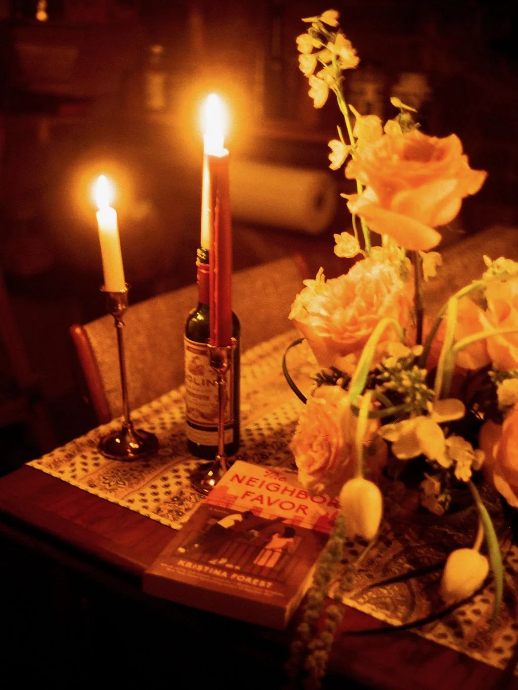 A dimly lit table with two lit candles in bottles, a bouquet of peach-colored flowers, and a book titled 'The Neighbor's Favor.'