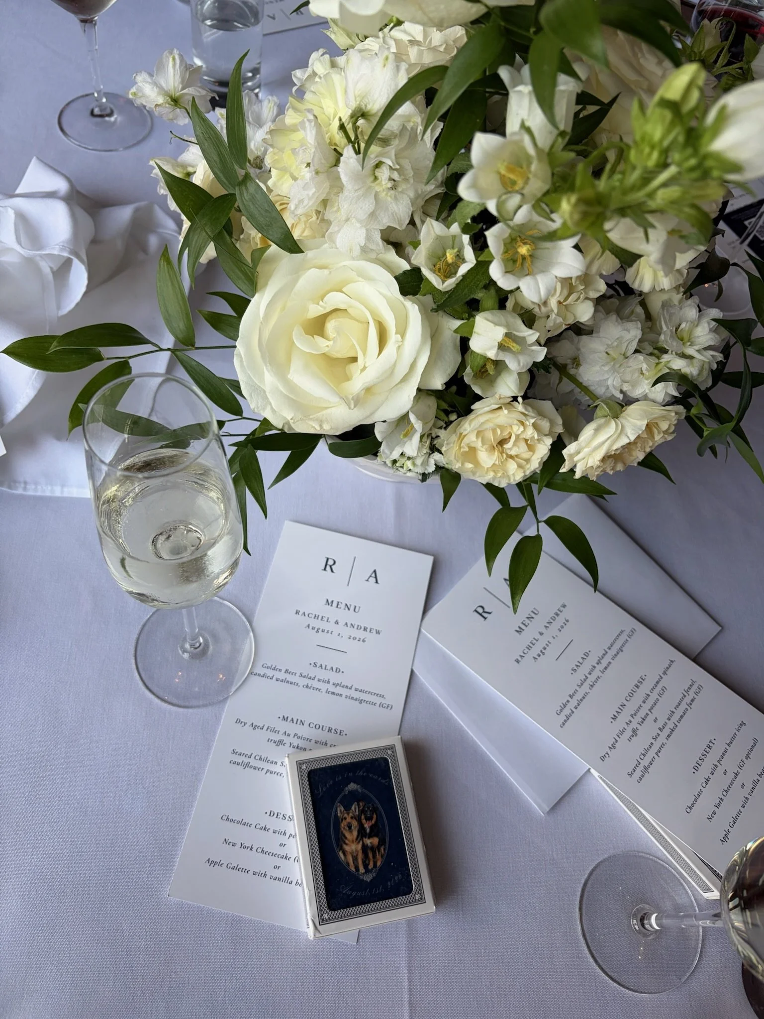 A wedding reception table decorated with white and cream flowers, green leaves, a wine glass, a water glass, and printed menu cards. There is a small boxed item with a dog illustration on the table.
