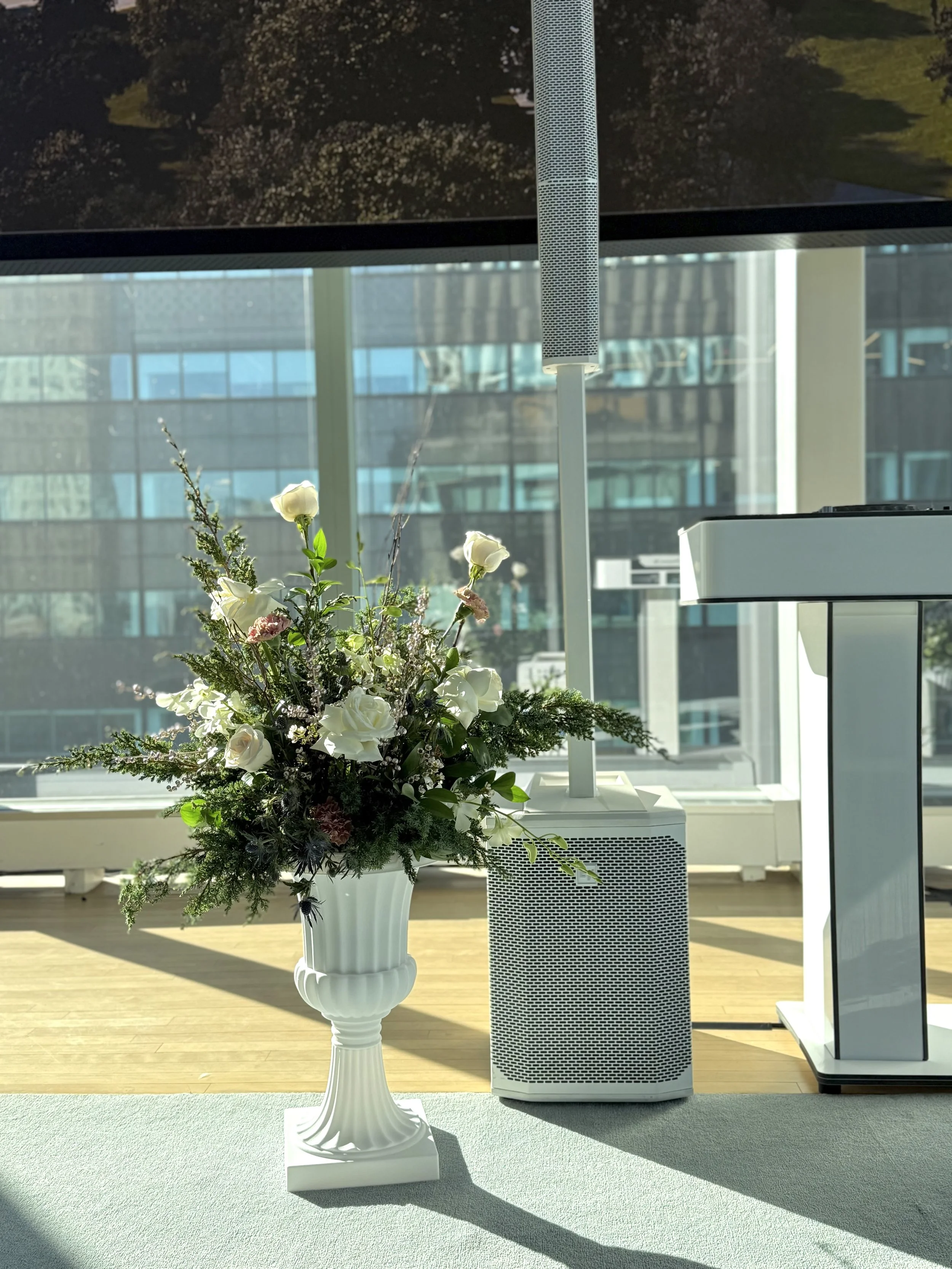 A white vase with a bouquet of white and pink flowers on a table, next to a white speaker and a large monitor with a cityscape view through windows in the background.