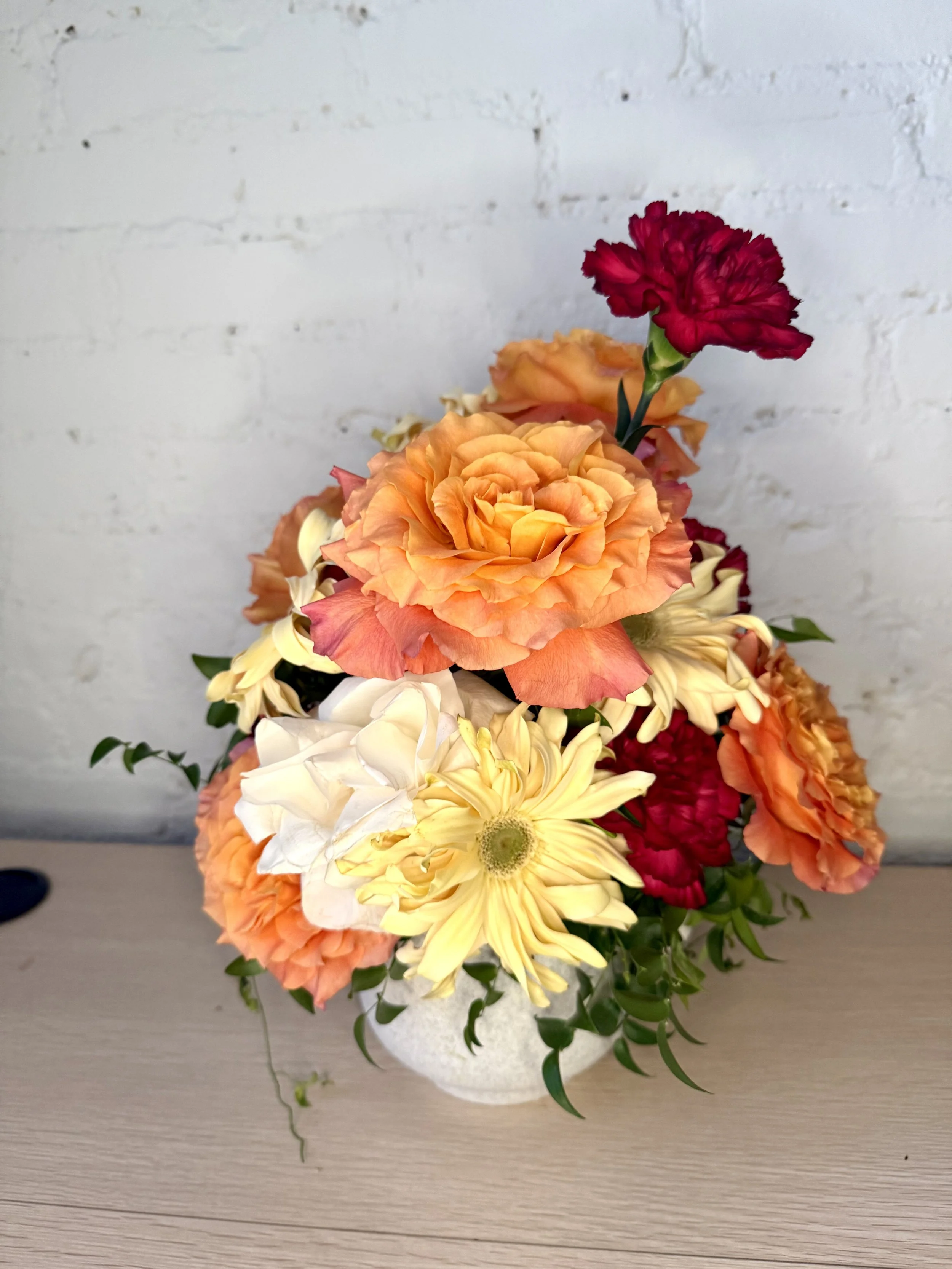 A bouquet of orange, yellow, white, and red flowers in a white vase on a wooden surface in front of a white brick wall.