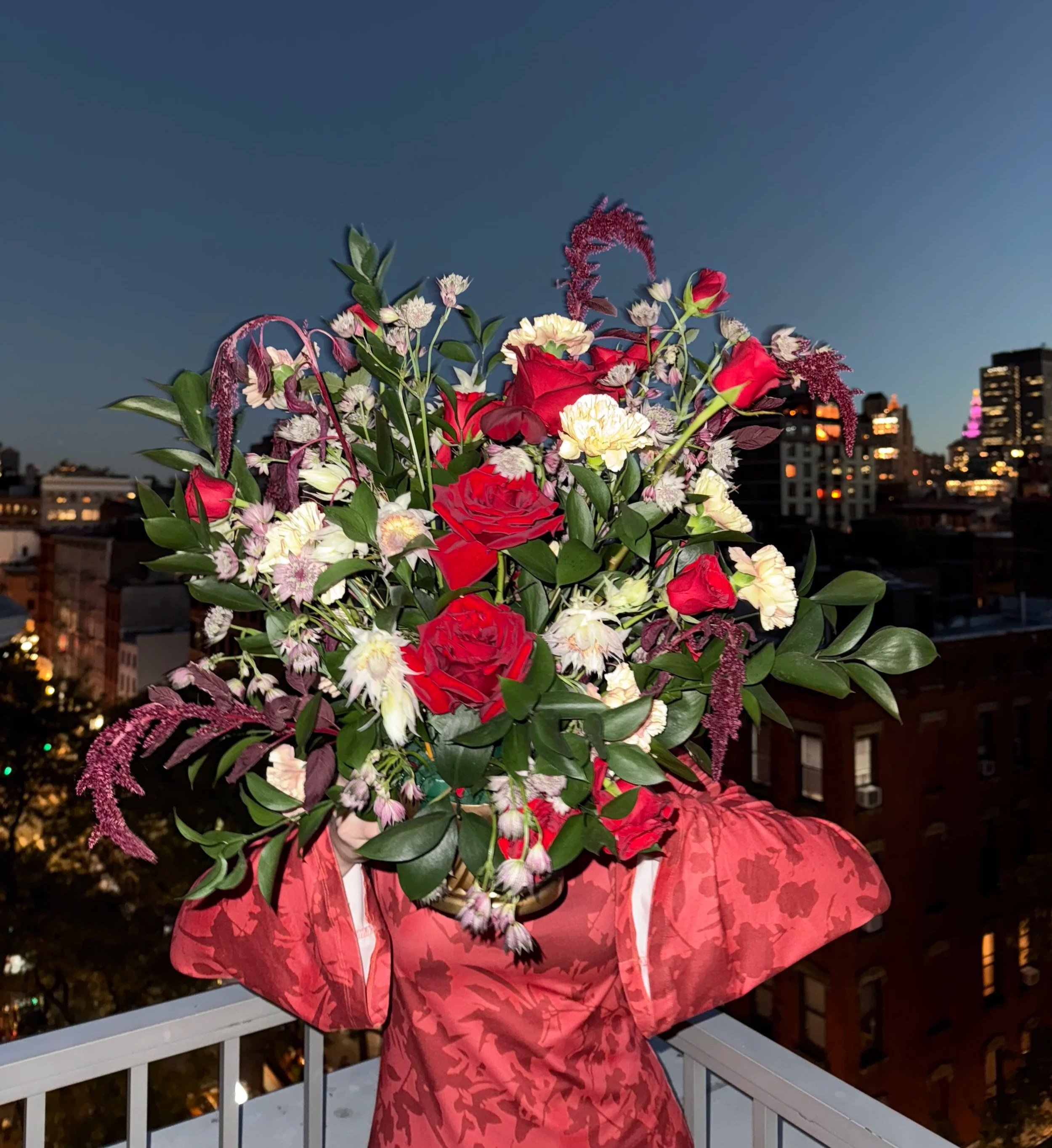 Person holding a large bouquet of colorful flowers on a balcony with city buildings and a darkening sky in the background.